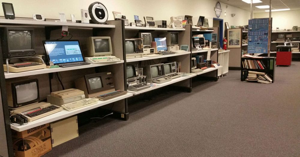 A row of vintage computers and monitors displayed on long shelves in a museum-like setting with carpet flooring.