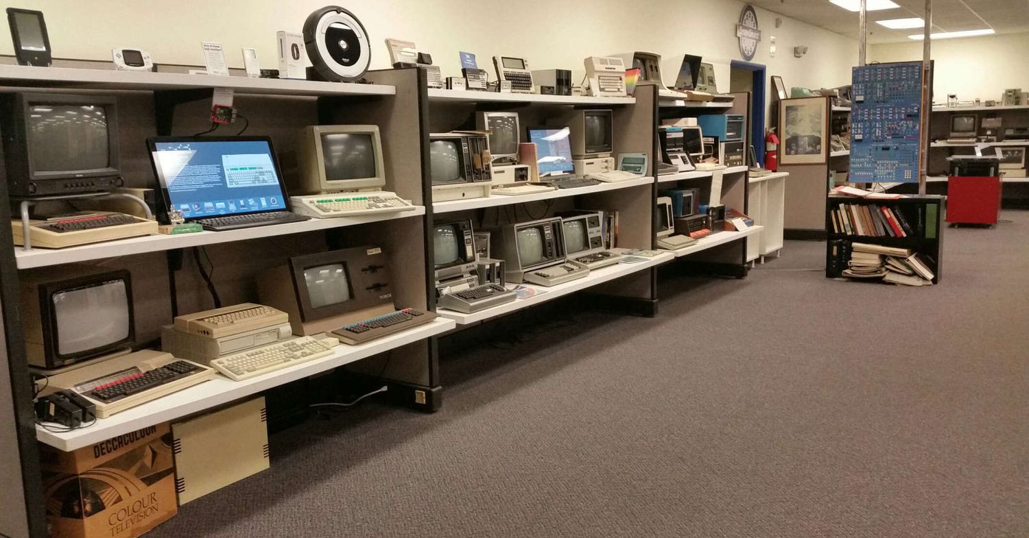 A row of vintage computers and monitors displayed on long shelves in a museum-like setting with carpet flooring.