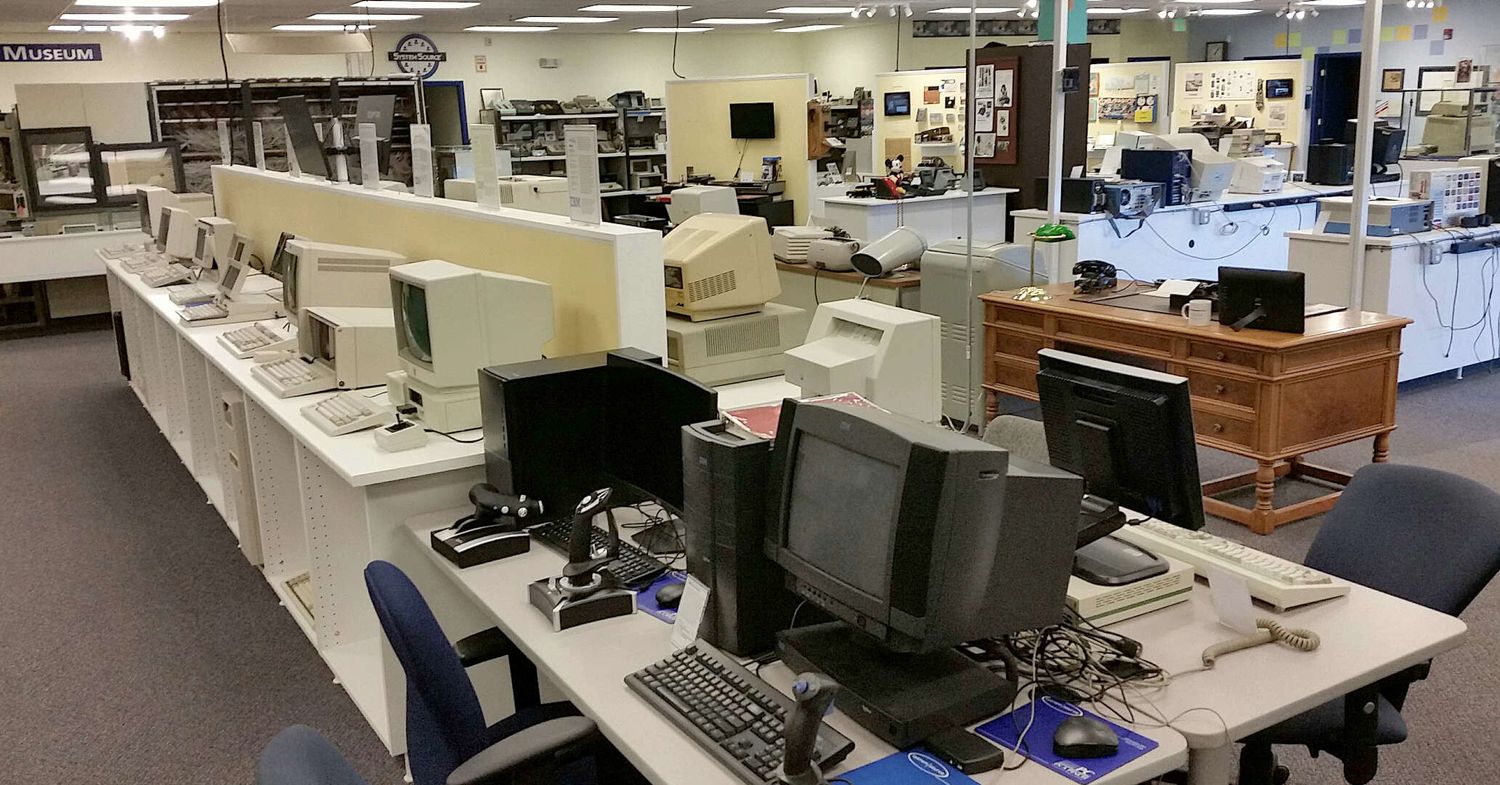 Computer workstations with vintage monitors and peripherals arranged in rows on tables in a large open office area.