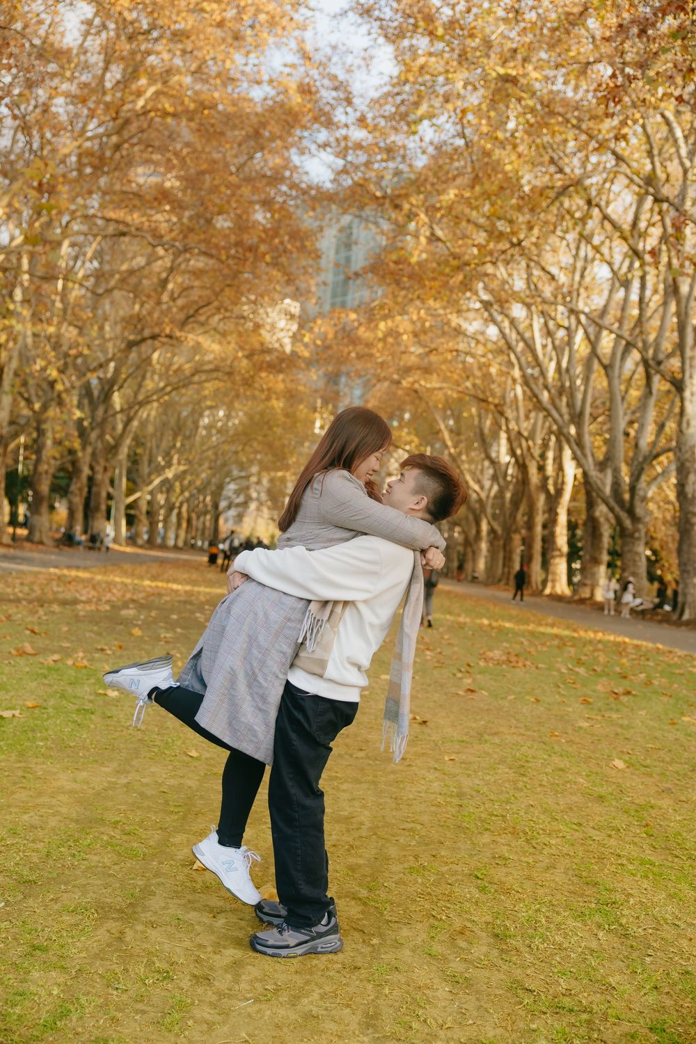 Couple sharing a playful embrace in a park surrounded by autumn trees with golden foliage.