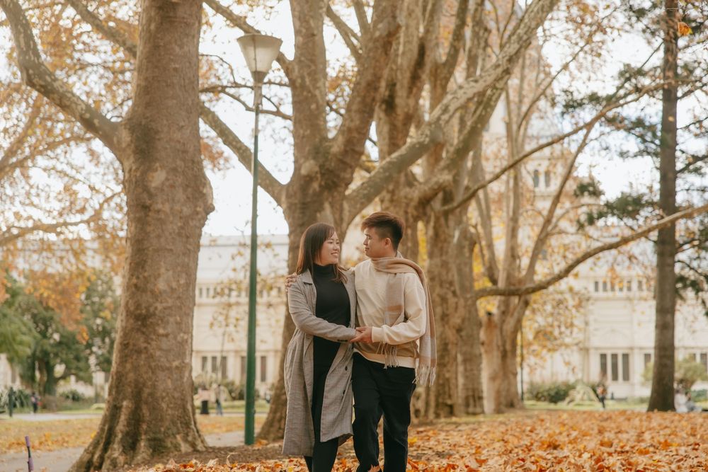 A couple shares an intimate moment among tall autumn trees with fallen leaves in a park.
