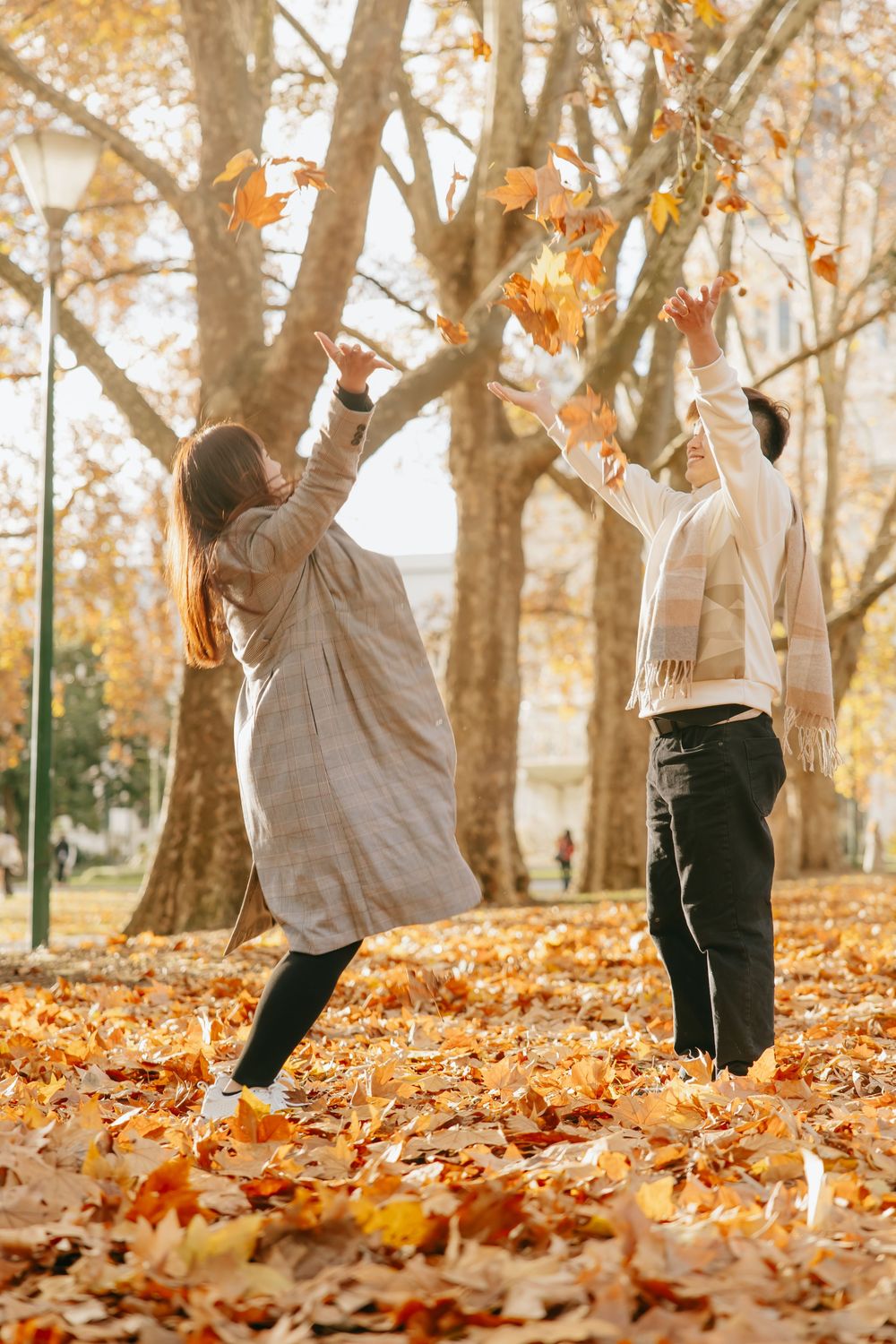 A person playfully jumping in a park filled with golden autumn leaves and tall trees.