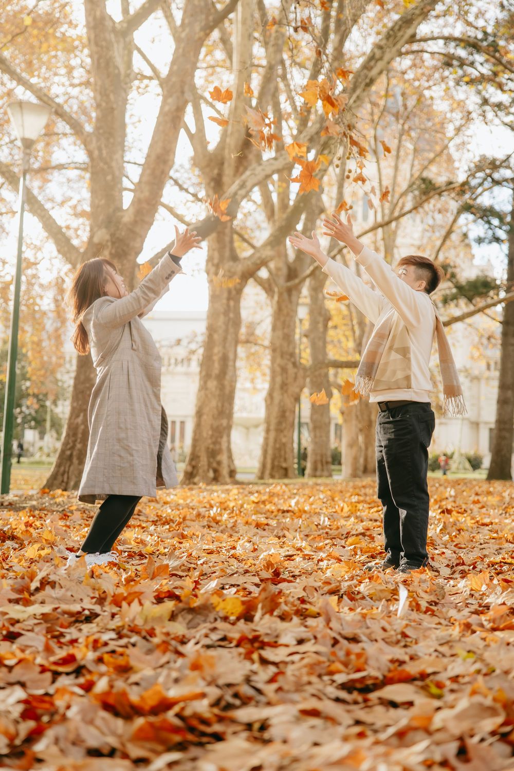 Joyful couple playing with falling autumn leaves in a park setting with trees overhead.
