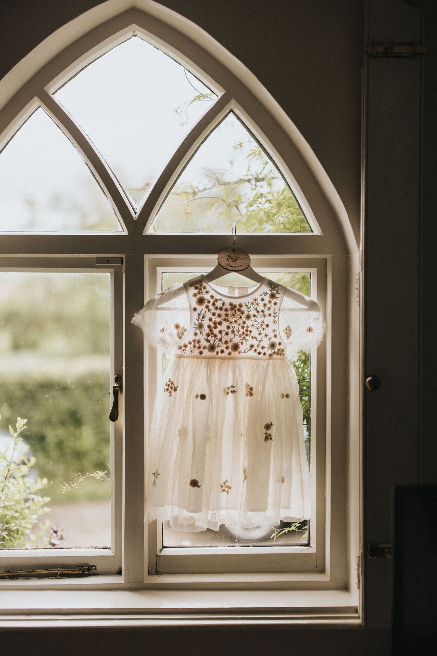 A delicate white embroidered dress hangs in an arched window frame with natural light streaming through - taken by Grimsby photographers.
