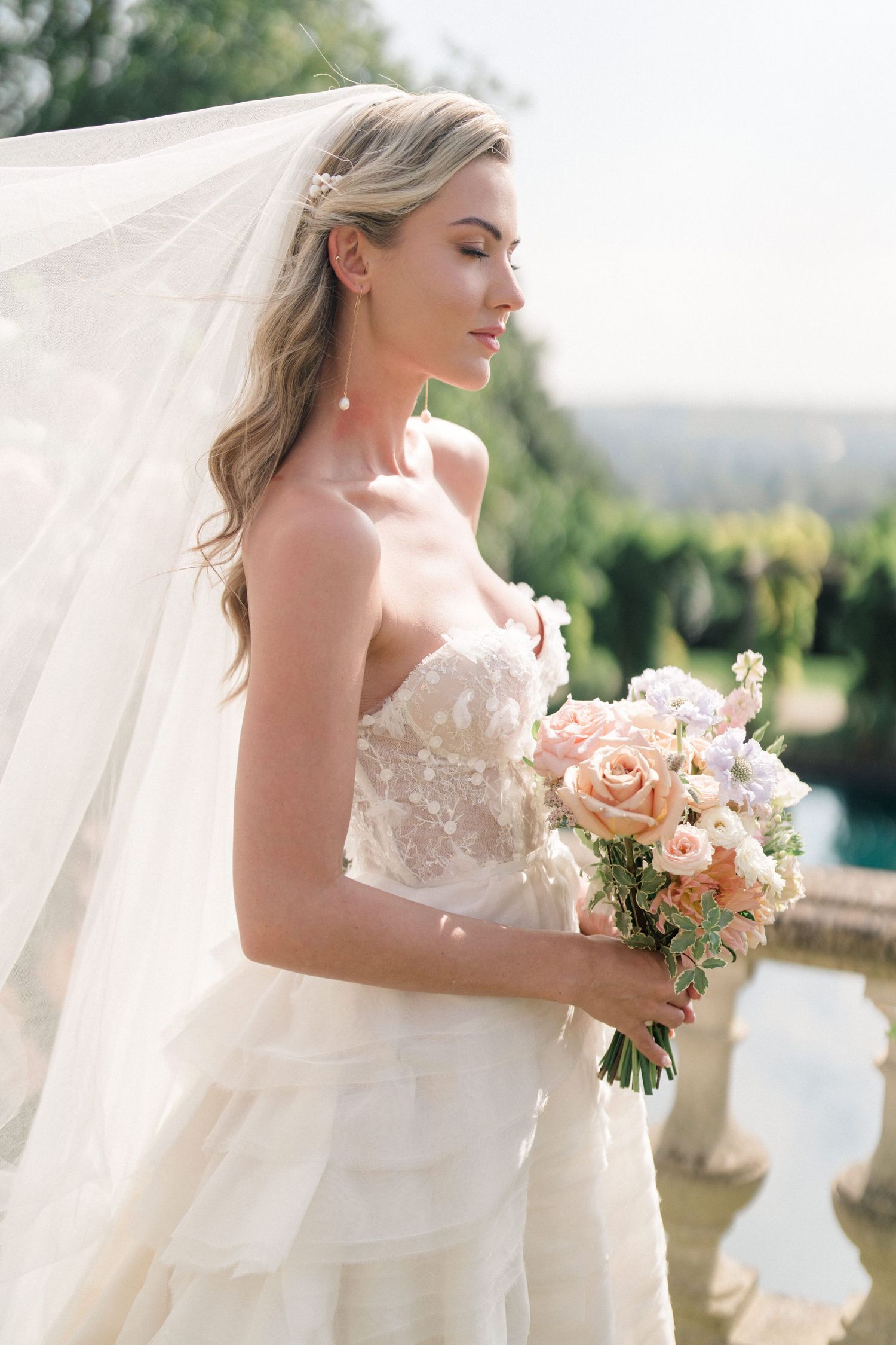 A bride in a strapless wedding gown holds a peach bouquet against an outdoor backdrop.