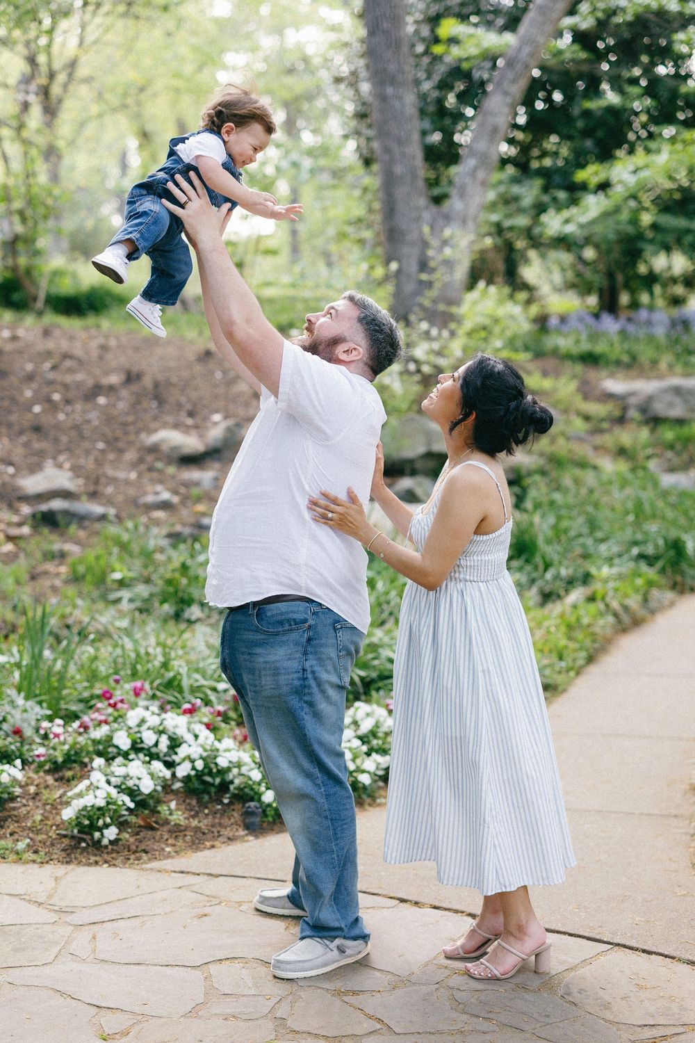 Playful family moment in a garden as they lift their baby up high while standing on a stone path.