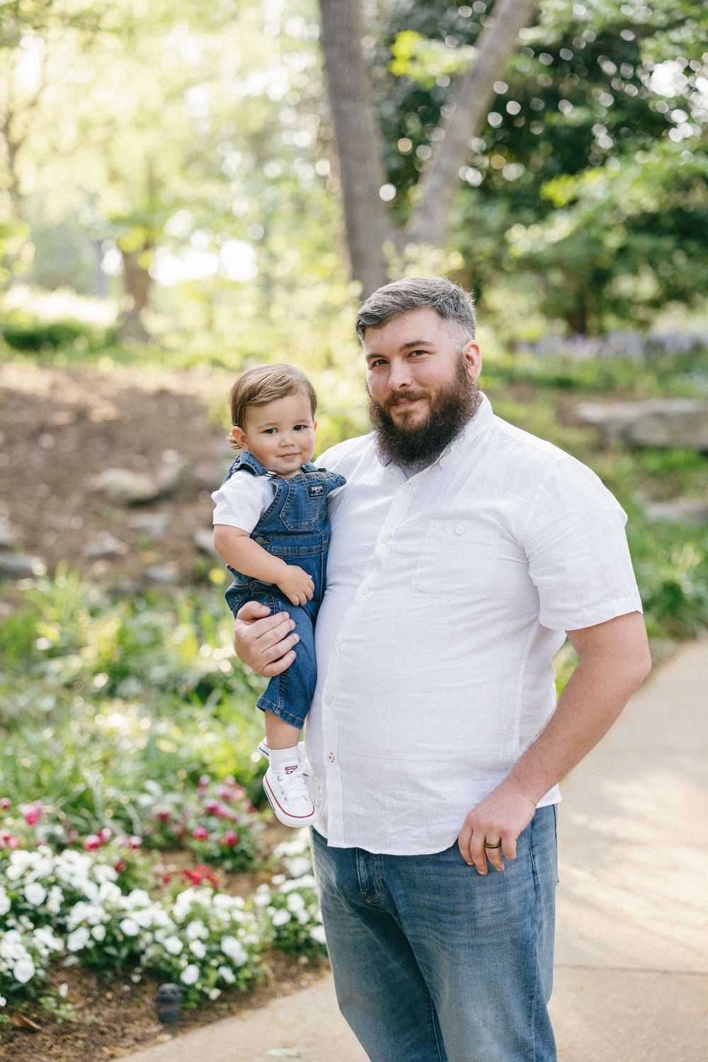 A person wearing a white shirt holds a small child in denim overalls on a garden pathway.