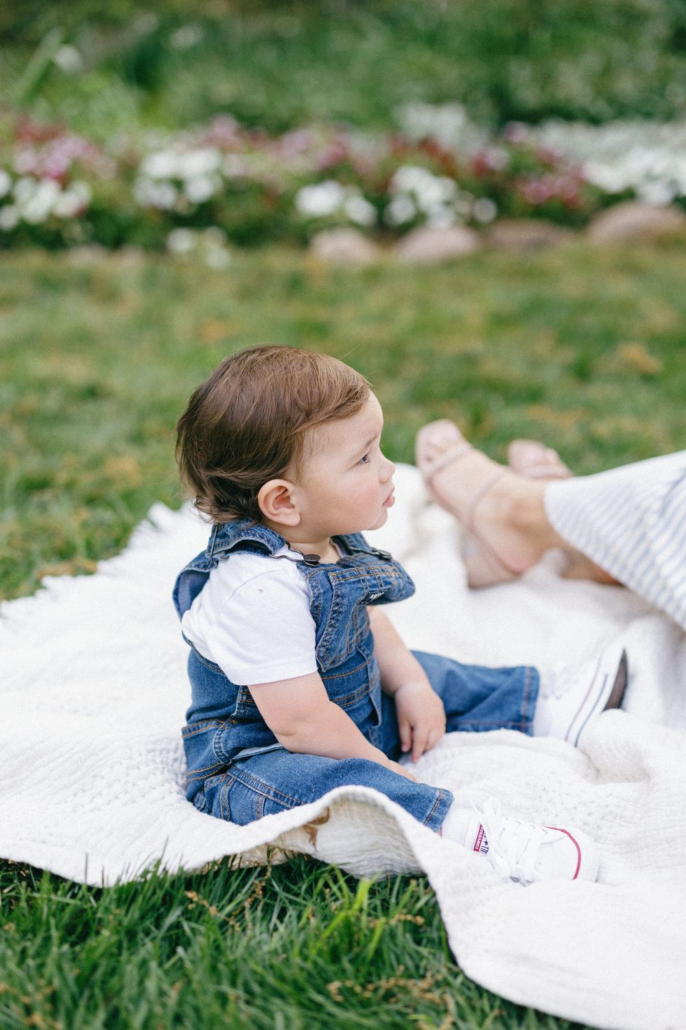 A baby wearing denim overalls lies on a white blanket in the grass during an outdoor photo session.