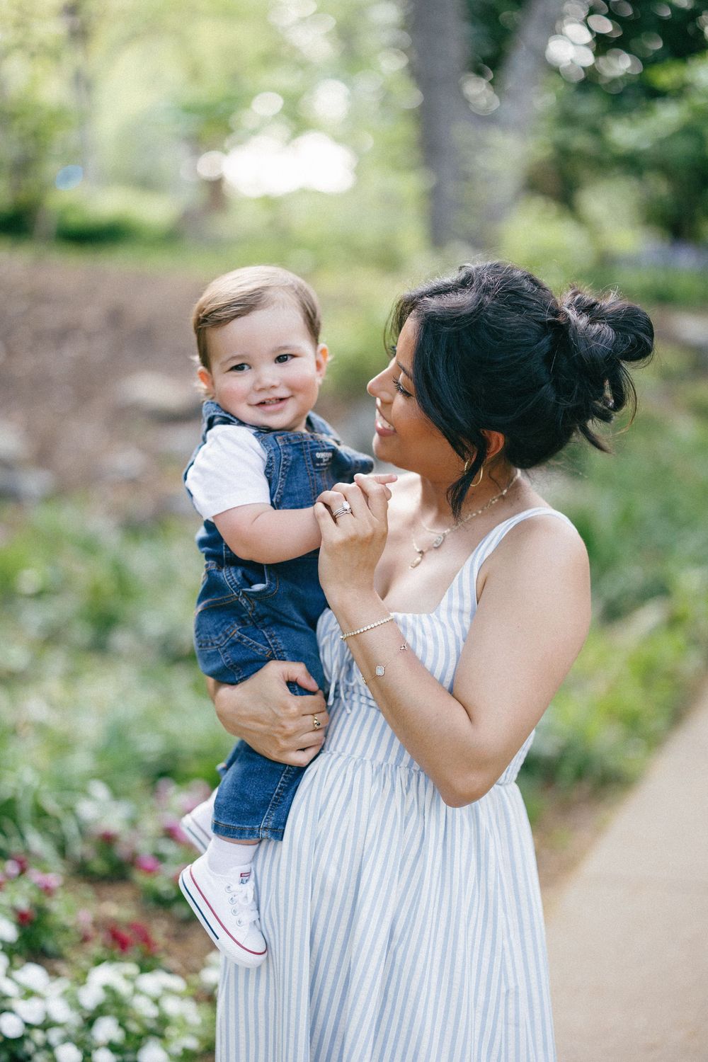 A tender outdoor moment between parent and child in a garden path surrounded by flowering bushes and greenery.