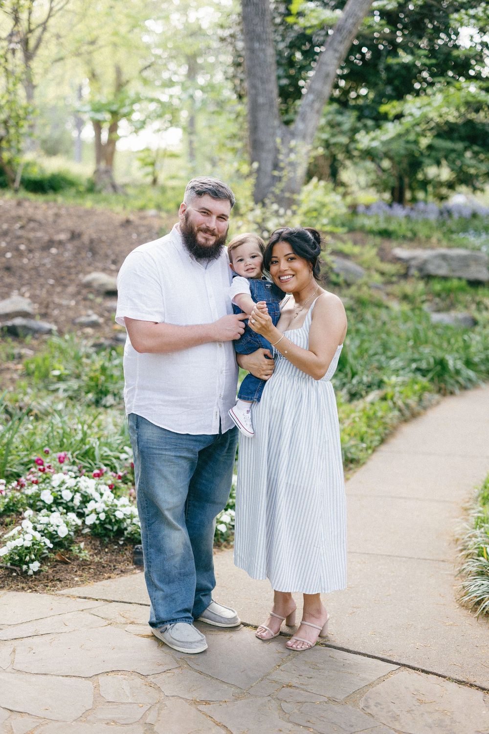A couple holds their toddler while standing on a garden path surrounded by blooming flowers and greenery.
