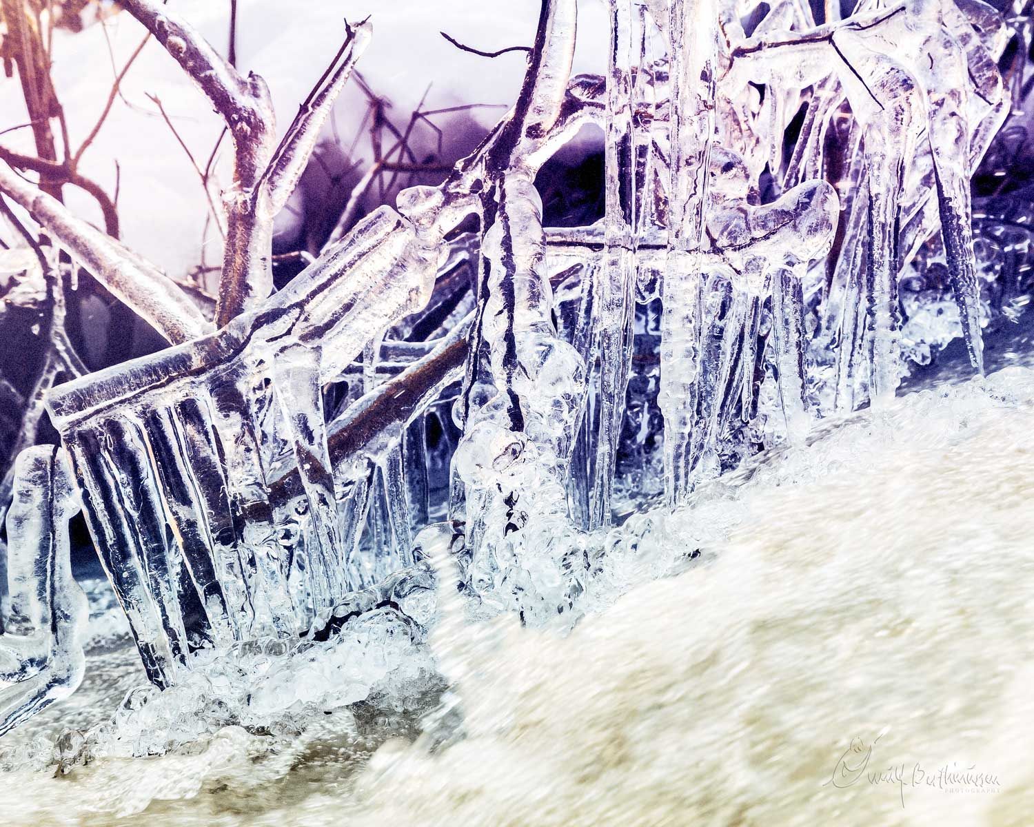 Frozen icicles hanging over rushing water create a dramatic winter scene with purple hues and crystalline formations.