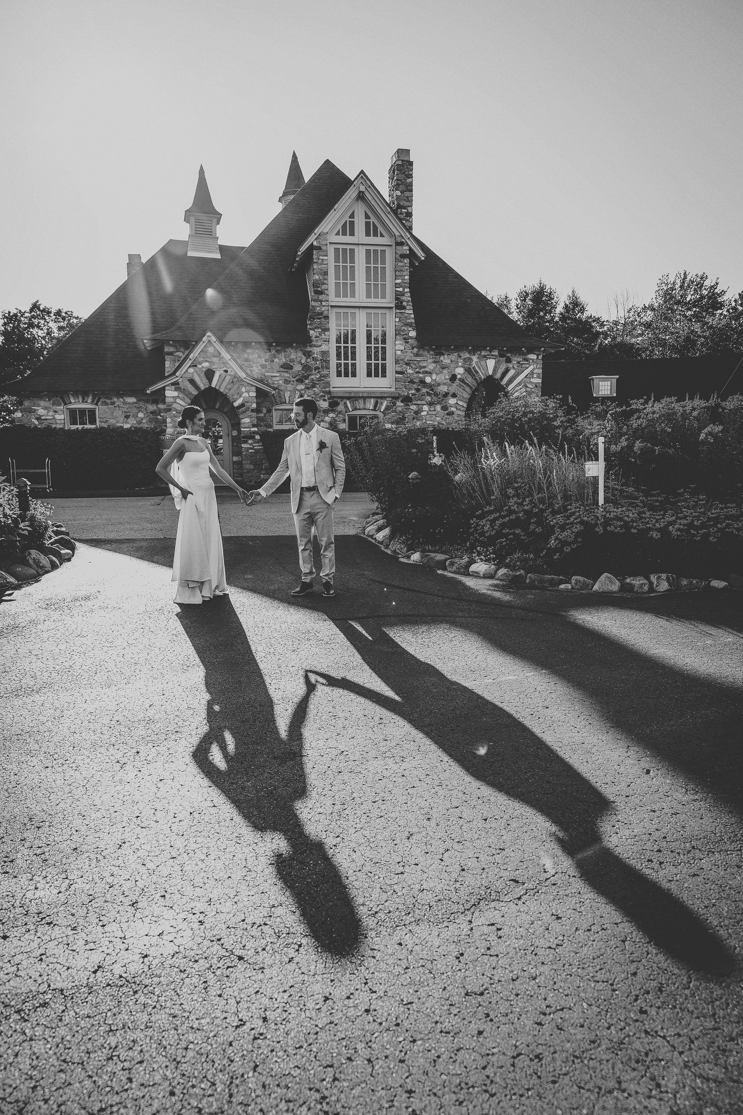 Black and white photo of a couple dancing in front of a stone cottage with dramatic long shadows cast on the ground.
