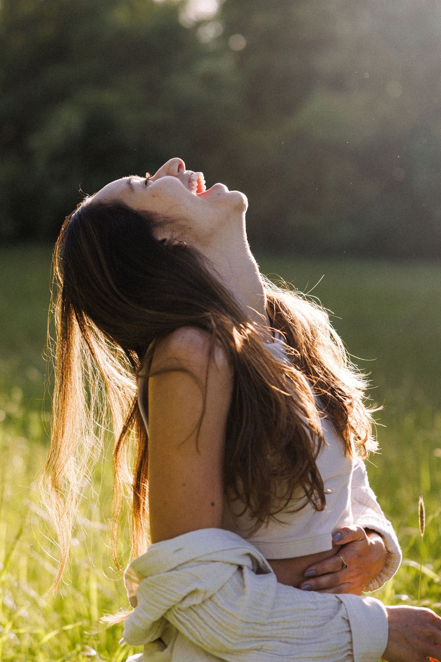 Person in white clothing sitting in sun-drenched meadow with head tilted back in joy.