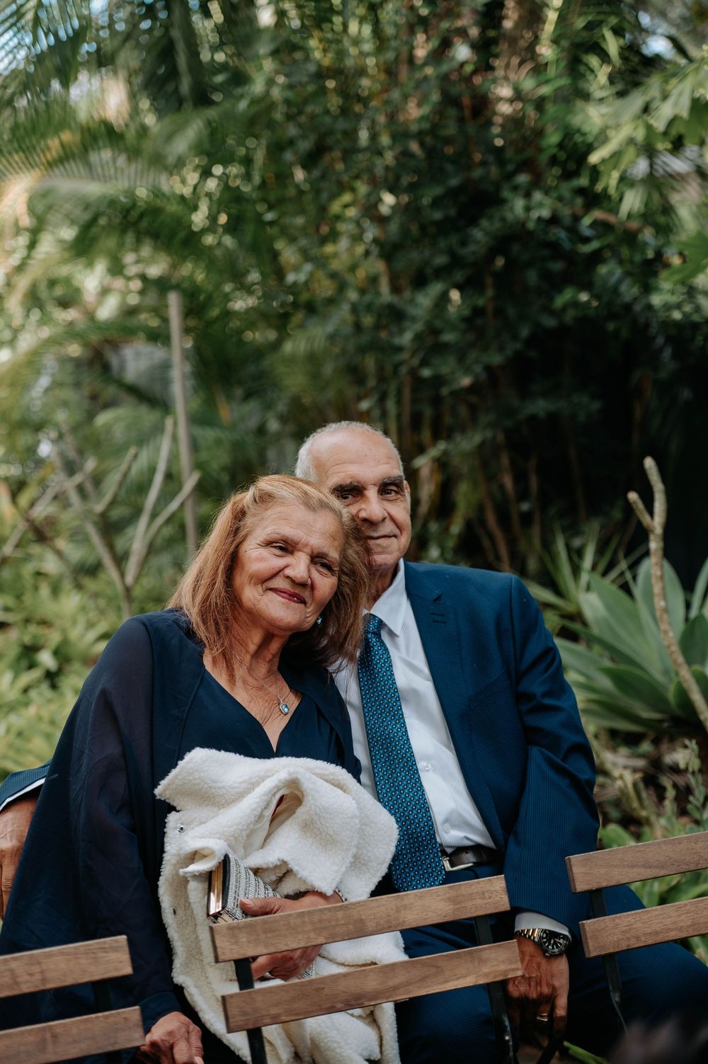 A couple sits together on a wooden bench surrounded by lush tropical plants and greenery.