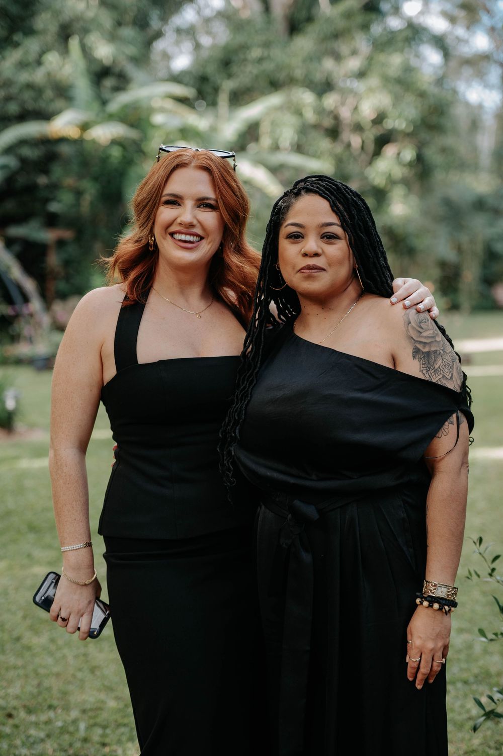 Two women stand together outdoors wearing elegant black formal dresses against a green natural background.