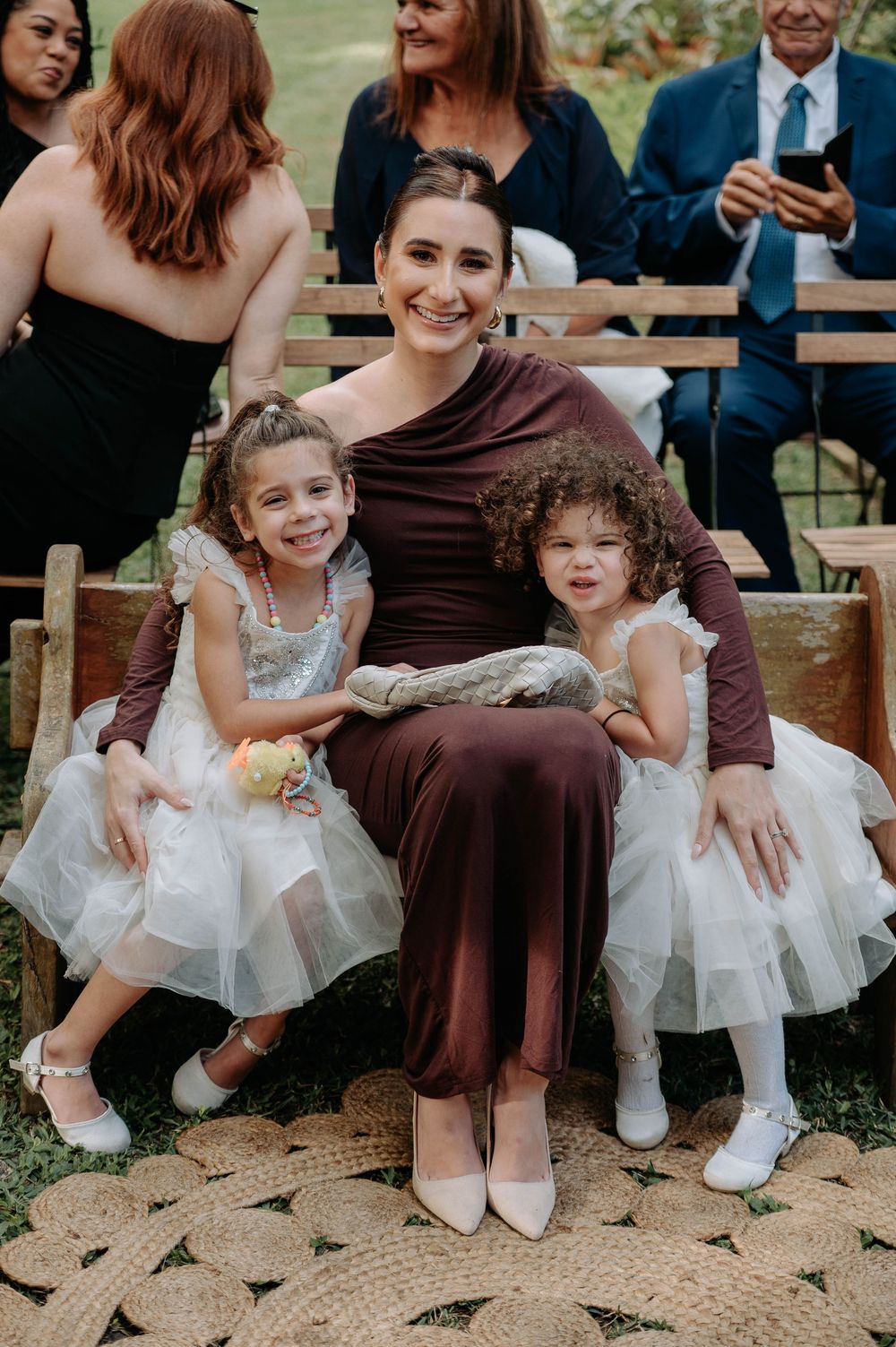 Woman in burgundy dress sits with two young flower girls in white tulle dresses at outdoor wedding event.