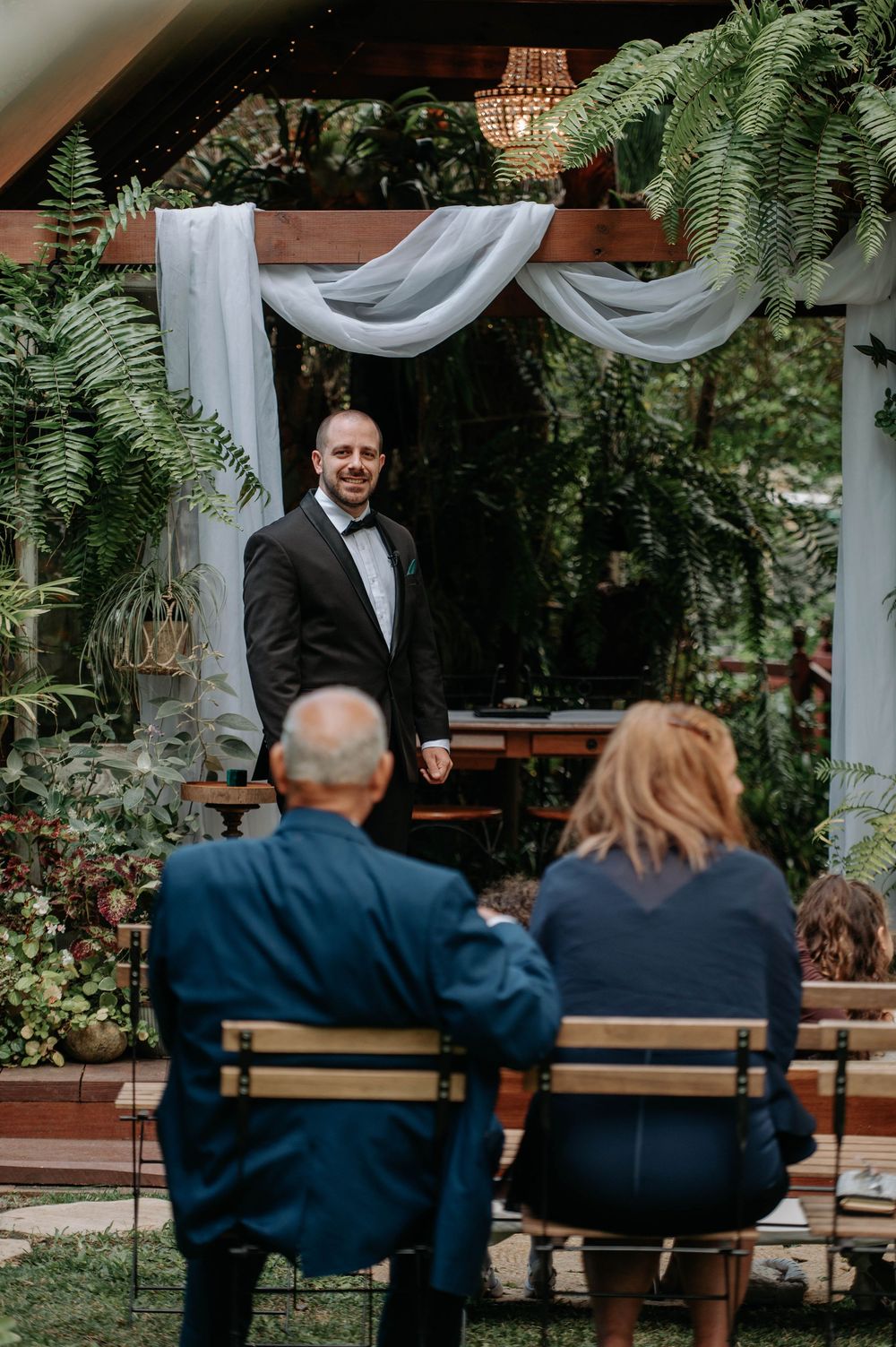 Outdoor garden wedding ceremony setup with guests seated on wooden benches under white fabric draping and lush greenery.