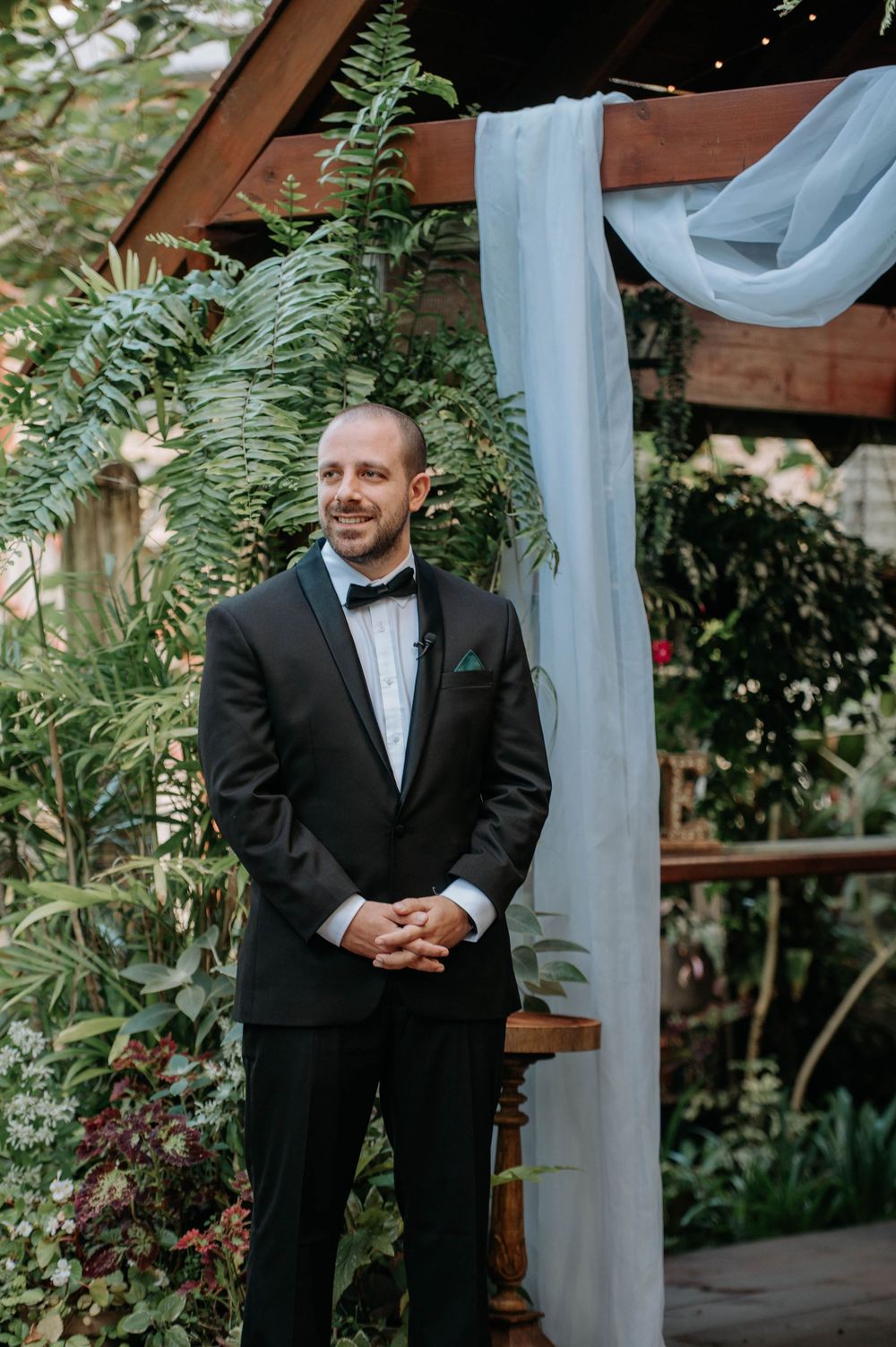 A groom in a black tuxedo stands beneath white draping and ferns at an outdoor wedding venue.