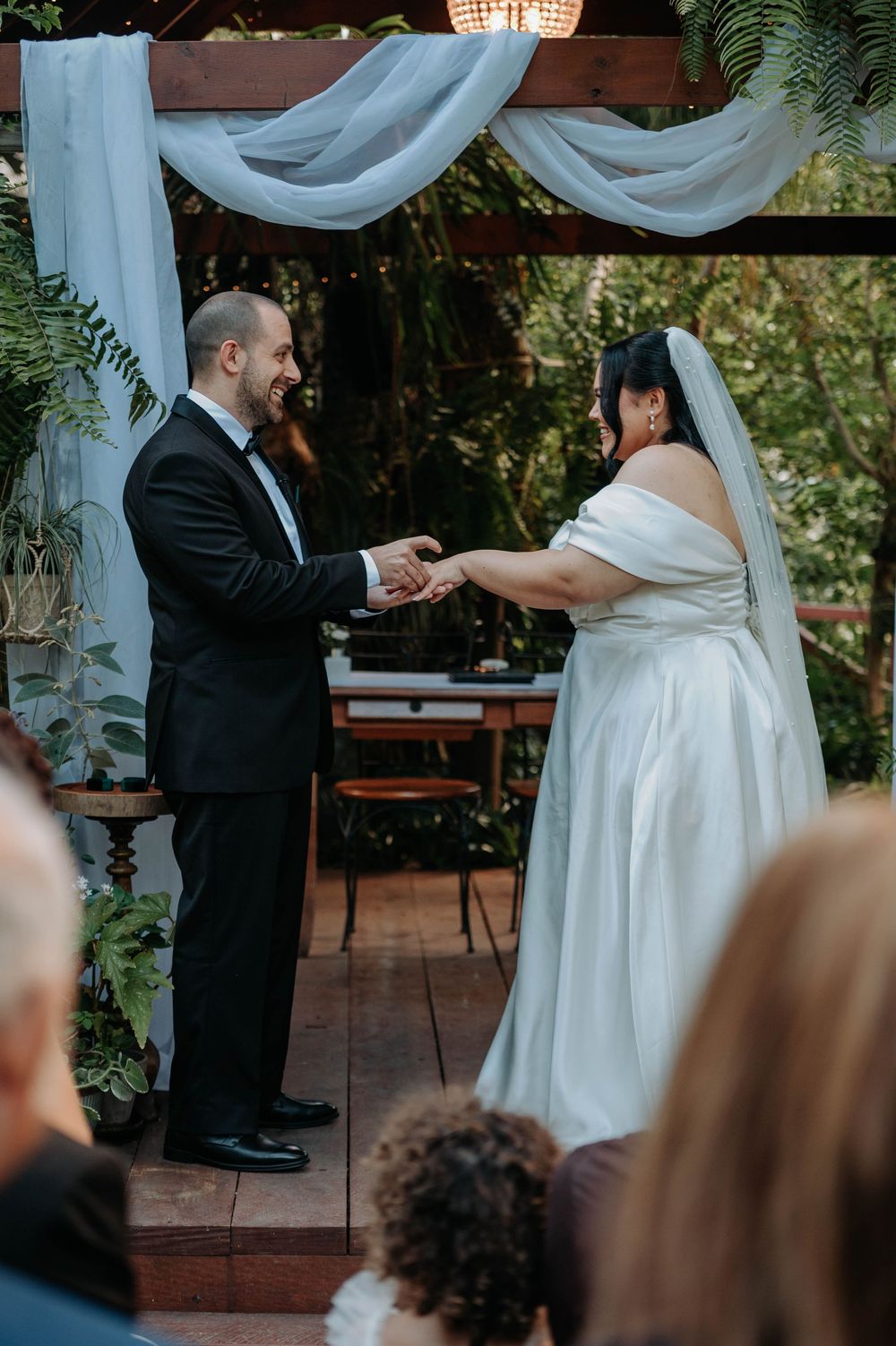 A couple exchanging rings during their outdoor wedding ceremony under draped white fabric and string lights.