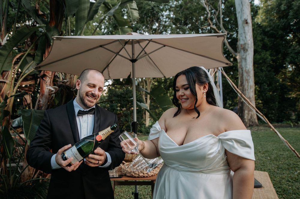 A joyful couple in wedding attire opens a champagne bottle under a beige umbrella in a garden setting.