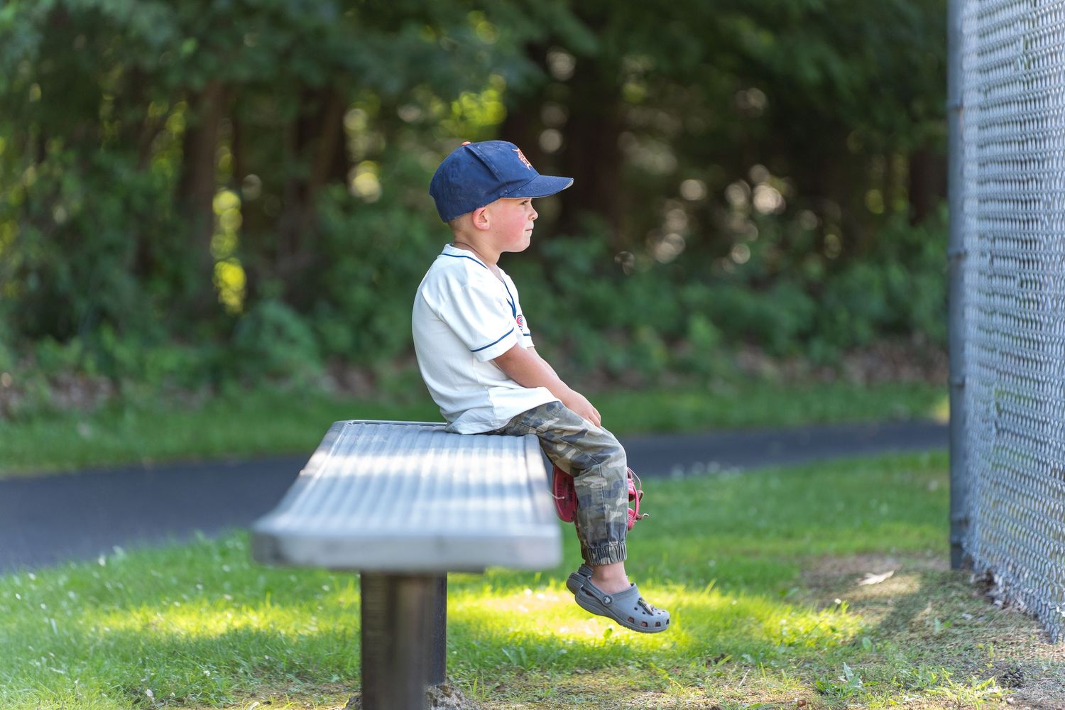 Person sits alone on a metal bench in a sunlit park setting with green grass and trees.