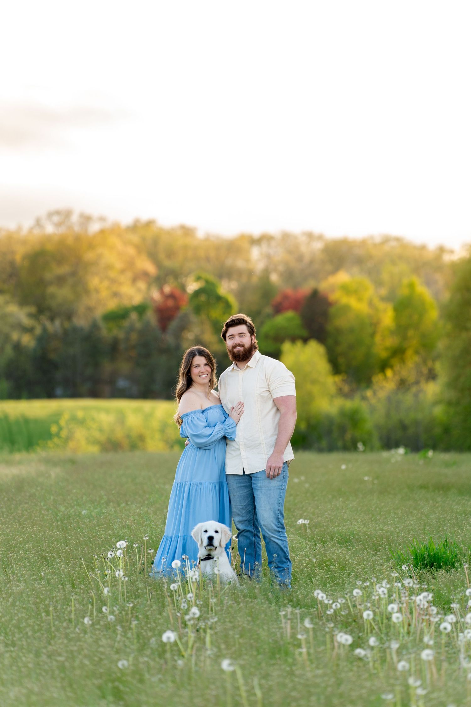A couple and their white dog stand together in a sunlit meadow filled with wildflowers during golden hour.