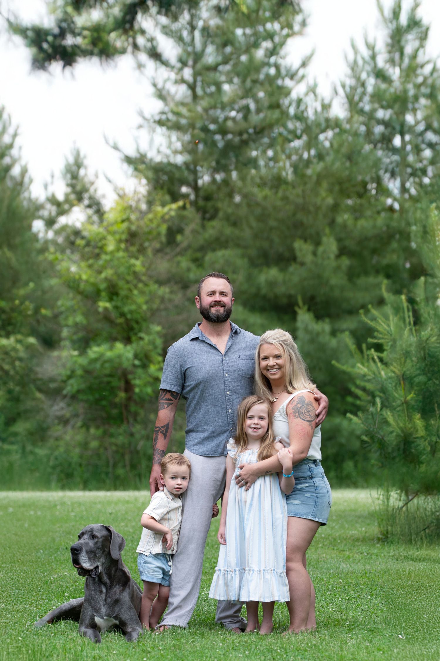Family poses together outdoors on a summer day with their black dog against a backdrop of evergreen trees.