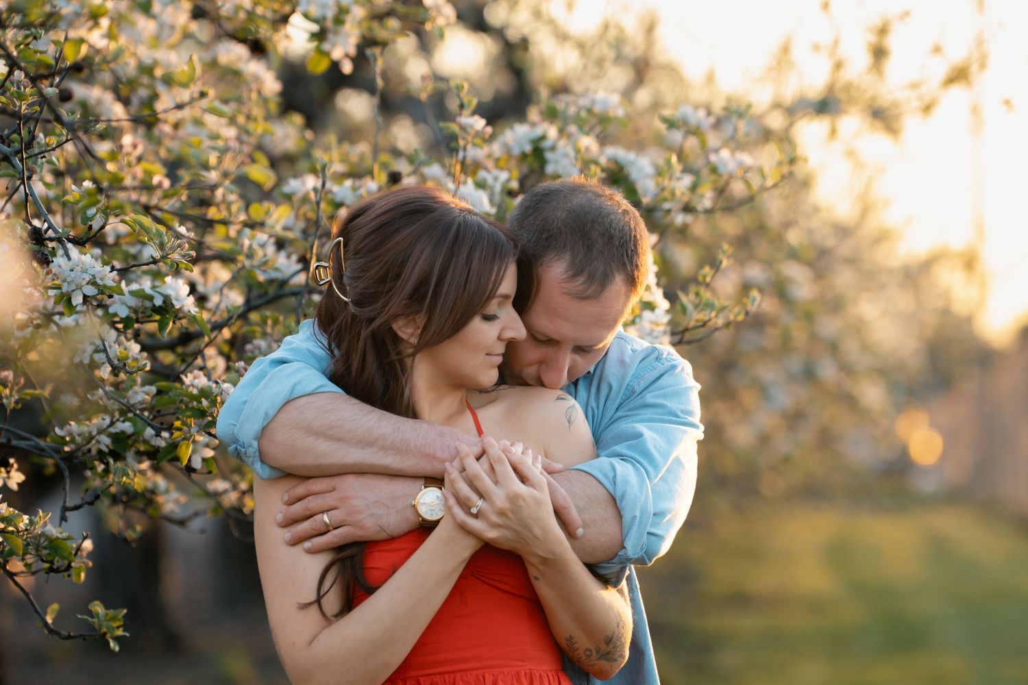 Two people embrace under blooming cherry blossom trees during a spring sunset in an orchard.