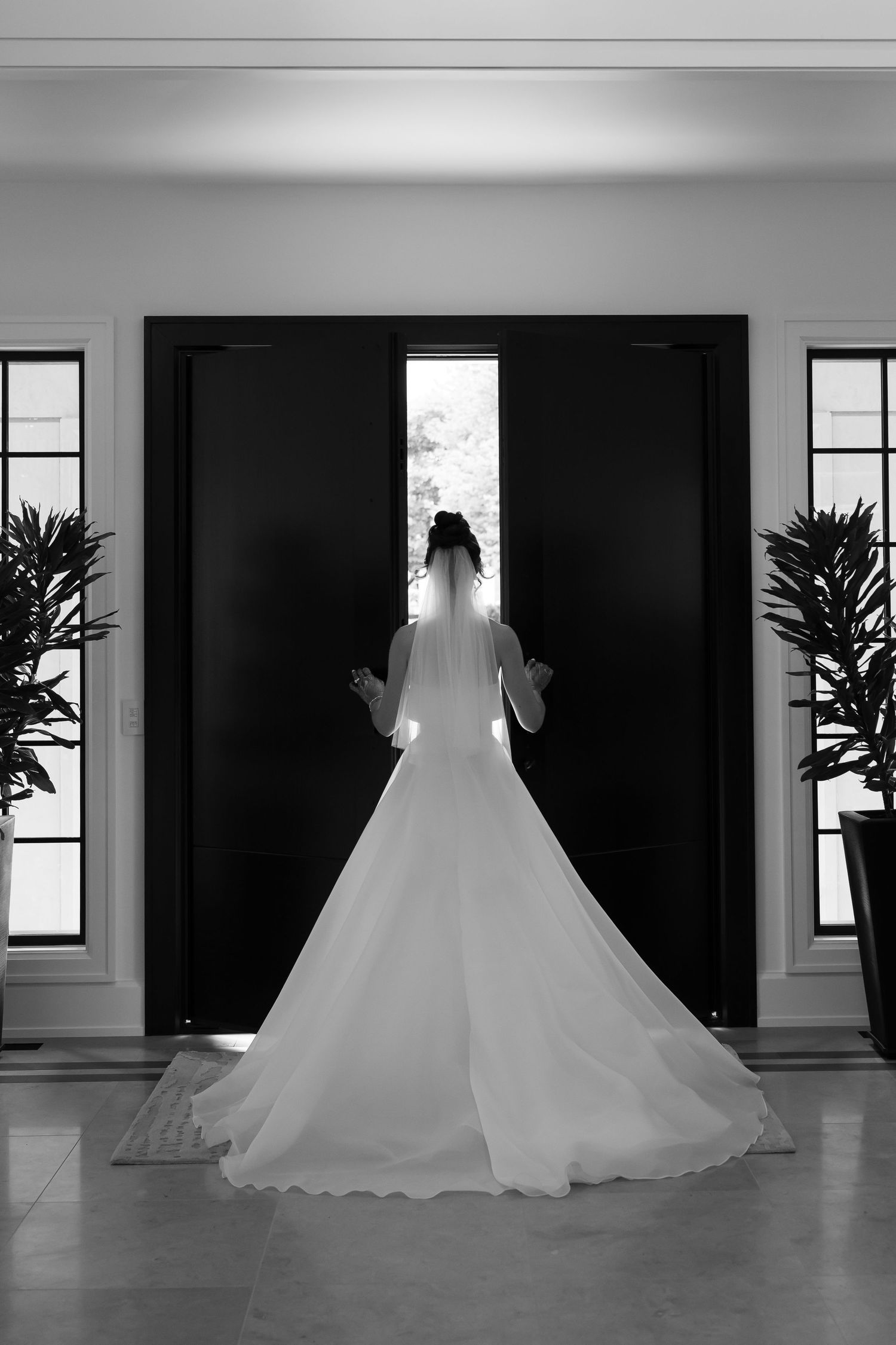 A silhouetted bride in a flowing white wedding gown poses in a minimalist room with tall windows and potted plants.
