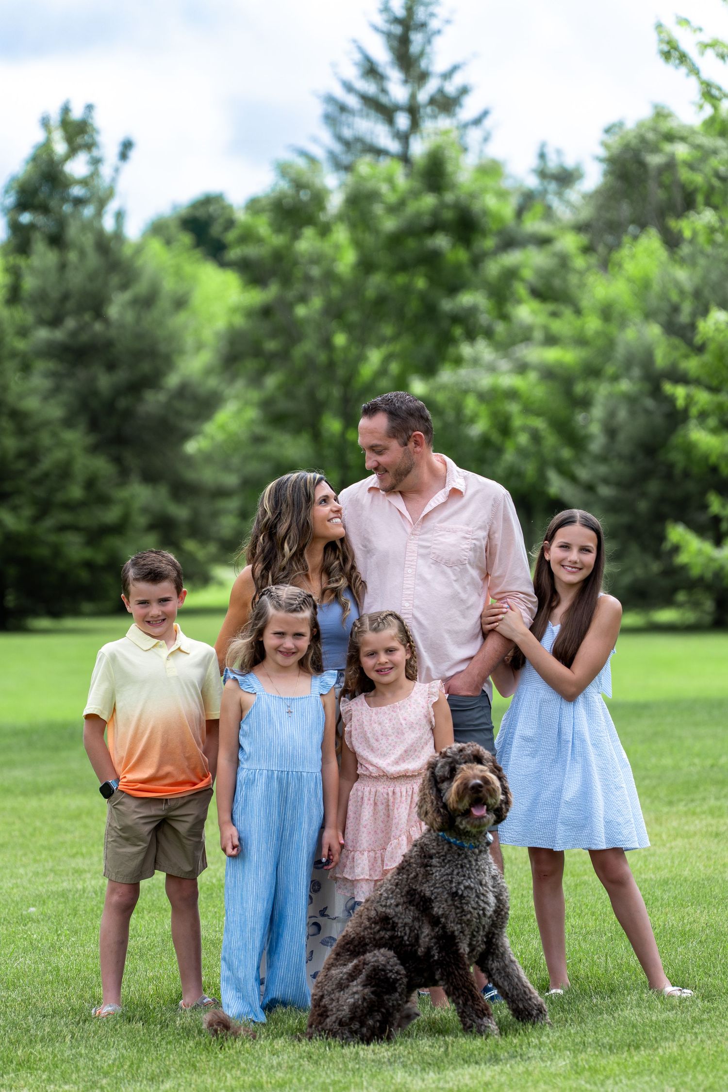 A family with three children stands together on a green lawn with their large brown curly-haired dog.
