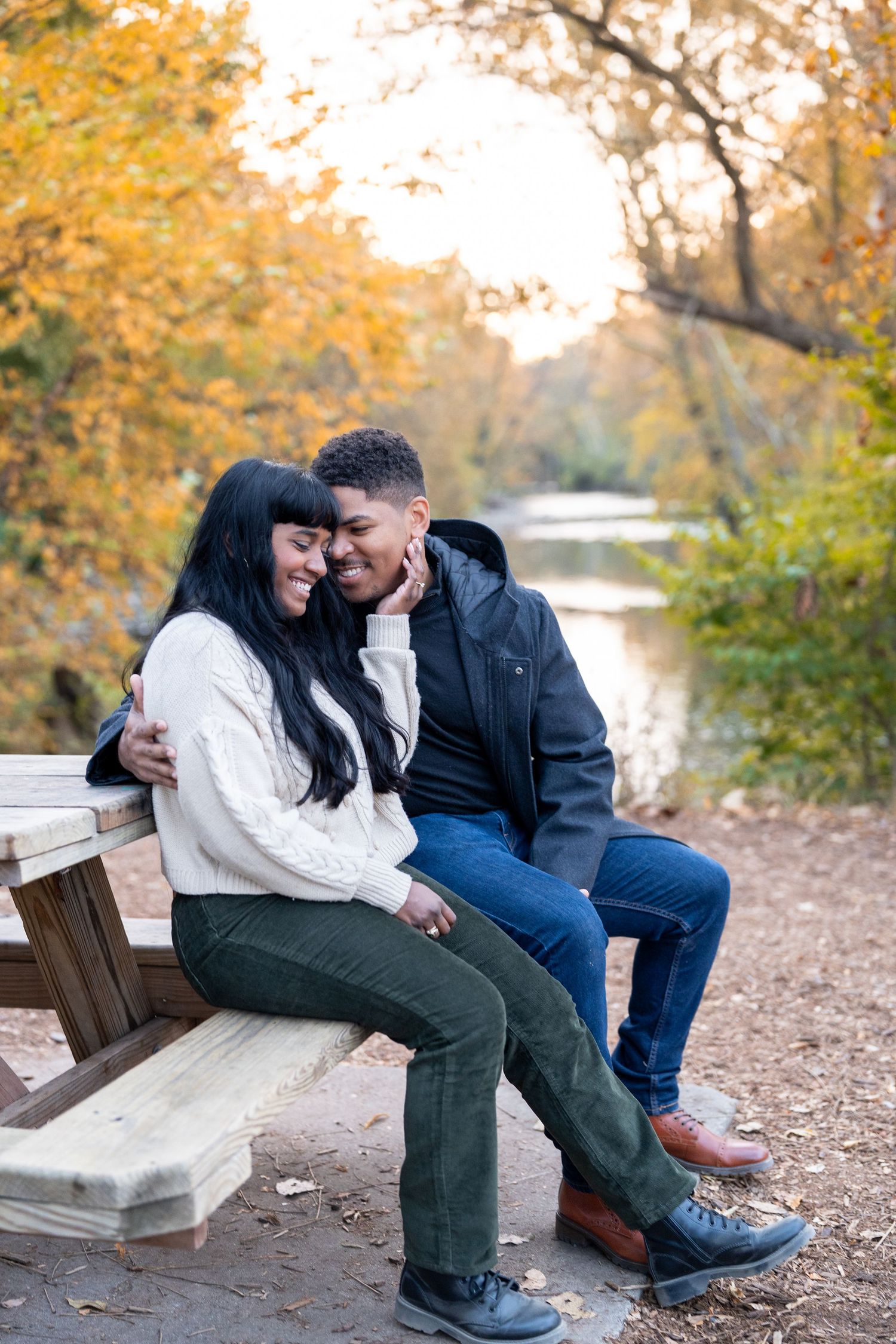 A couple shares a joyful moment together on a bench by a river during autumn with golden foliage in the background.