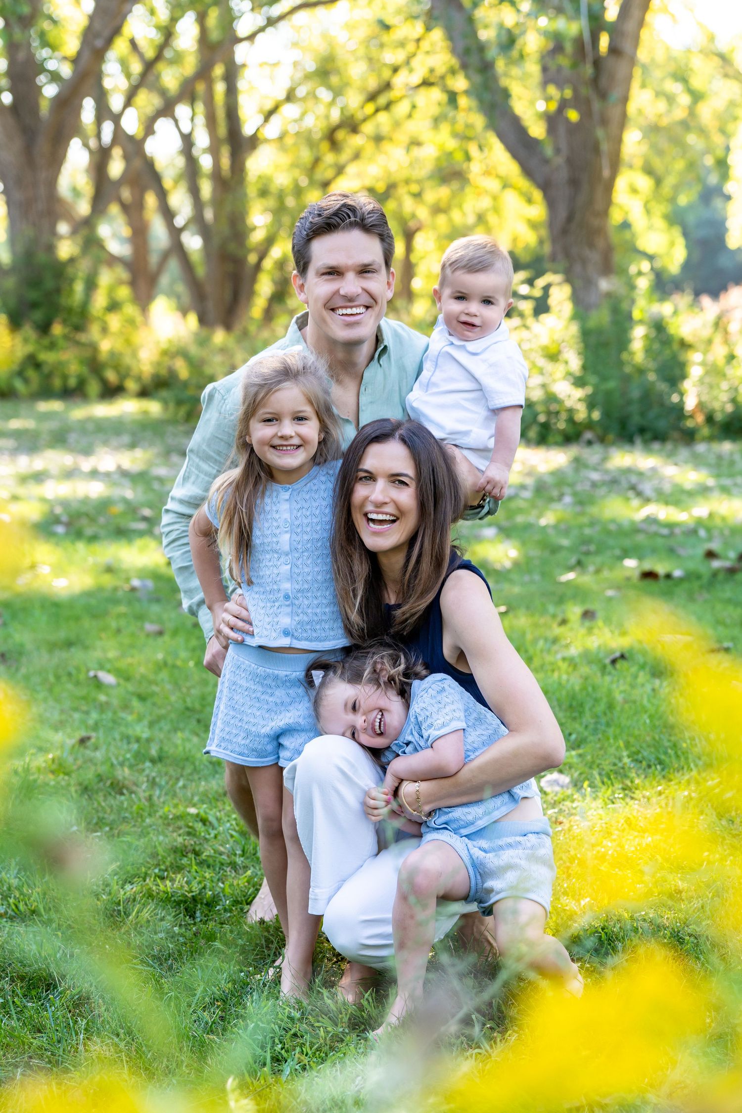 A family shares joyful moments together while posing in a sunny meadow with yellow wildflowers in the foreground.