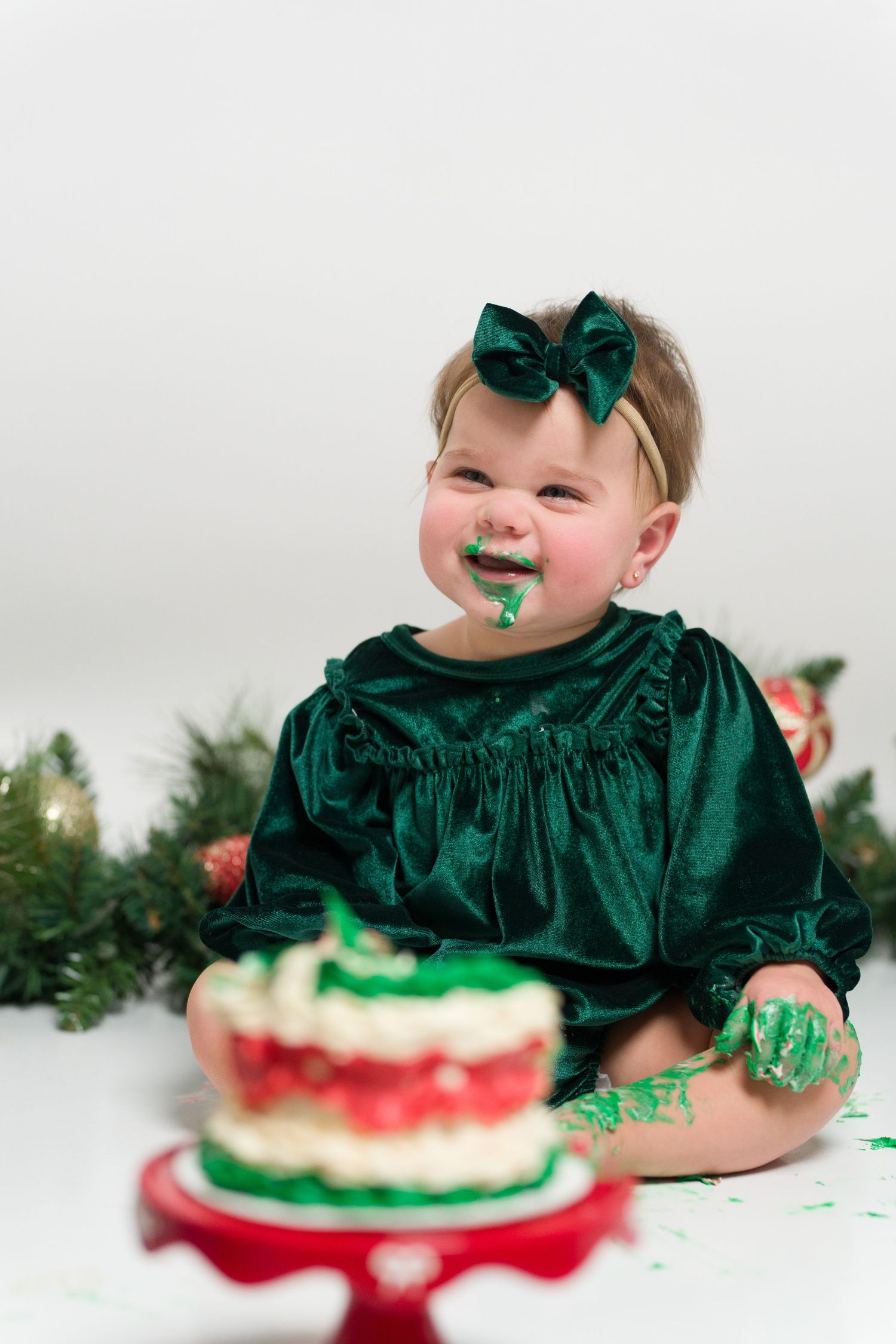 Baby in emerald green velvet dress smashes and plays with cake during festive Christmas cake smash photo session.