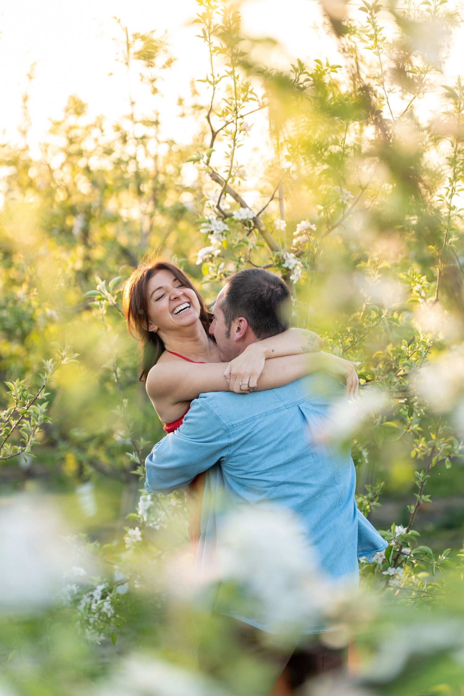 A joyful couple embraces in a sun-dappled spring orchard with white blossoms and golden light filtering through.