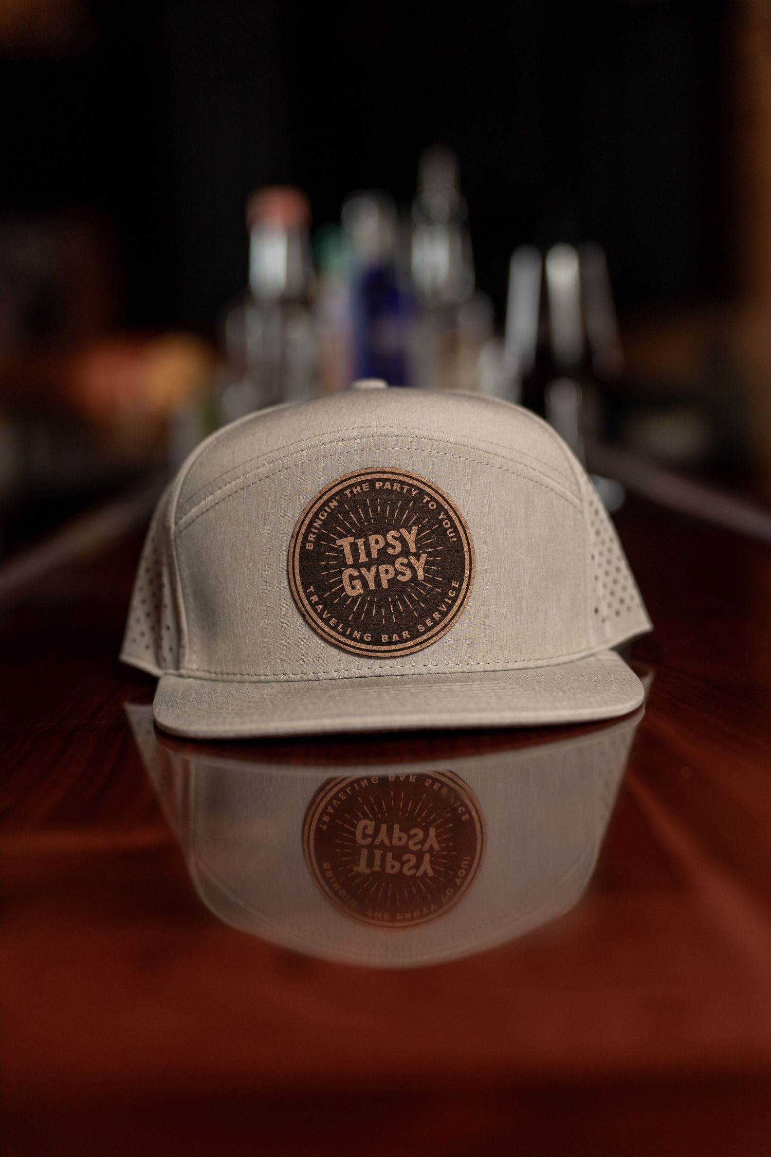 White baseball cap with brown Tipsy Gypsy patch logo resting on reflective wooden surface in dark bar setting.