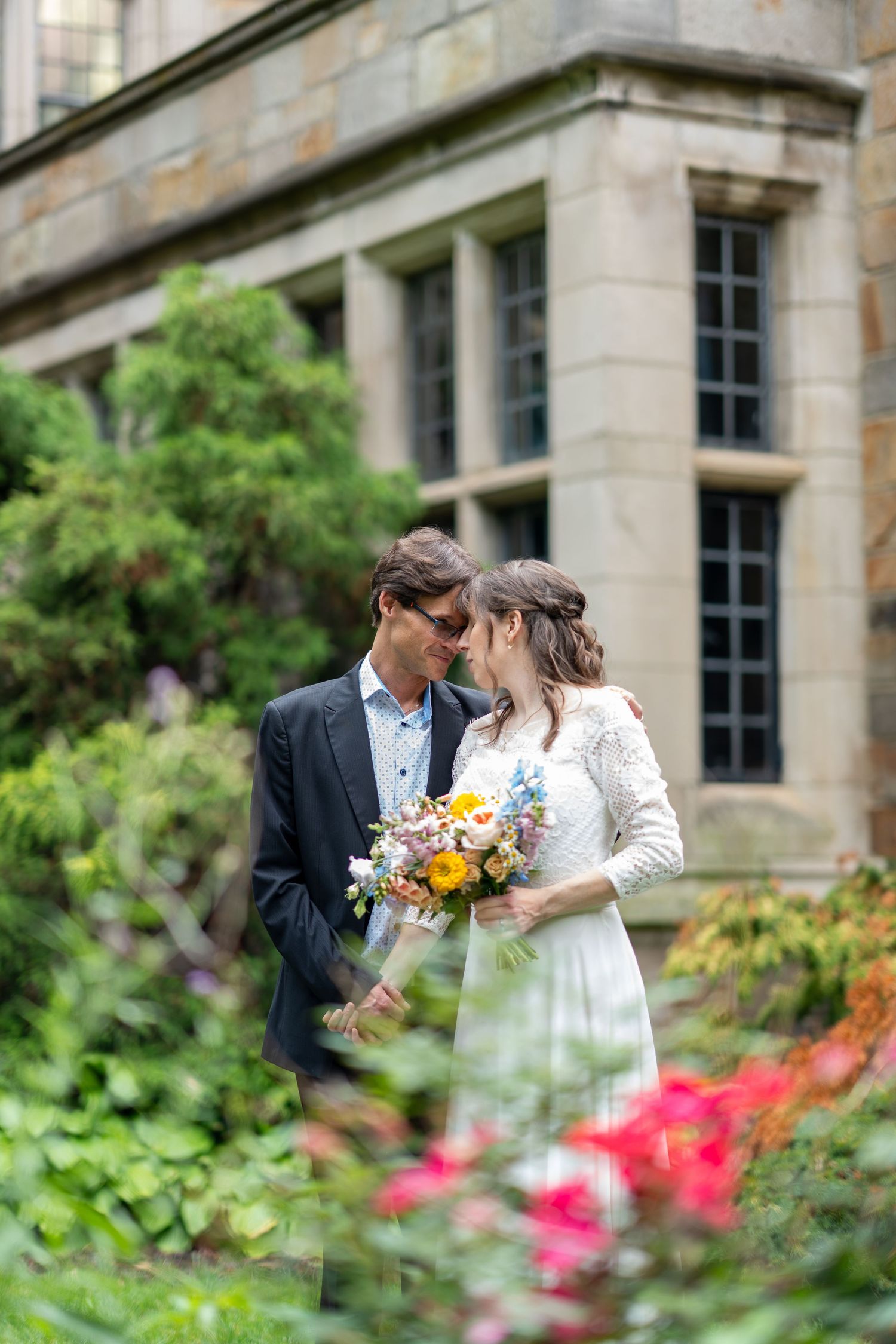 A couple shares an intimate moment in a lush garden setting with stone architecture and colorful flowers in the foreground.