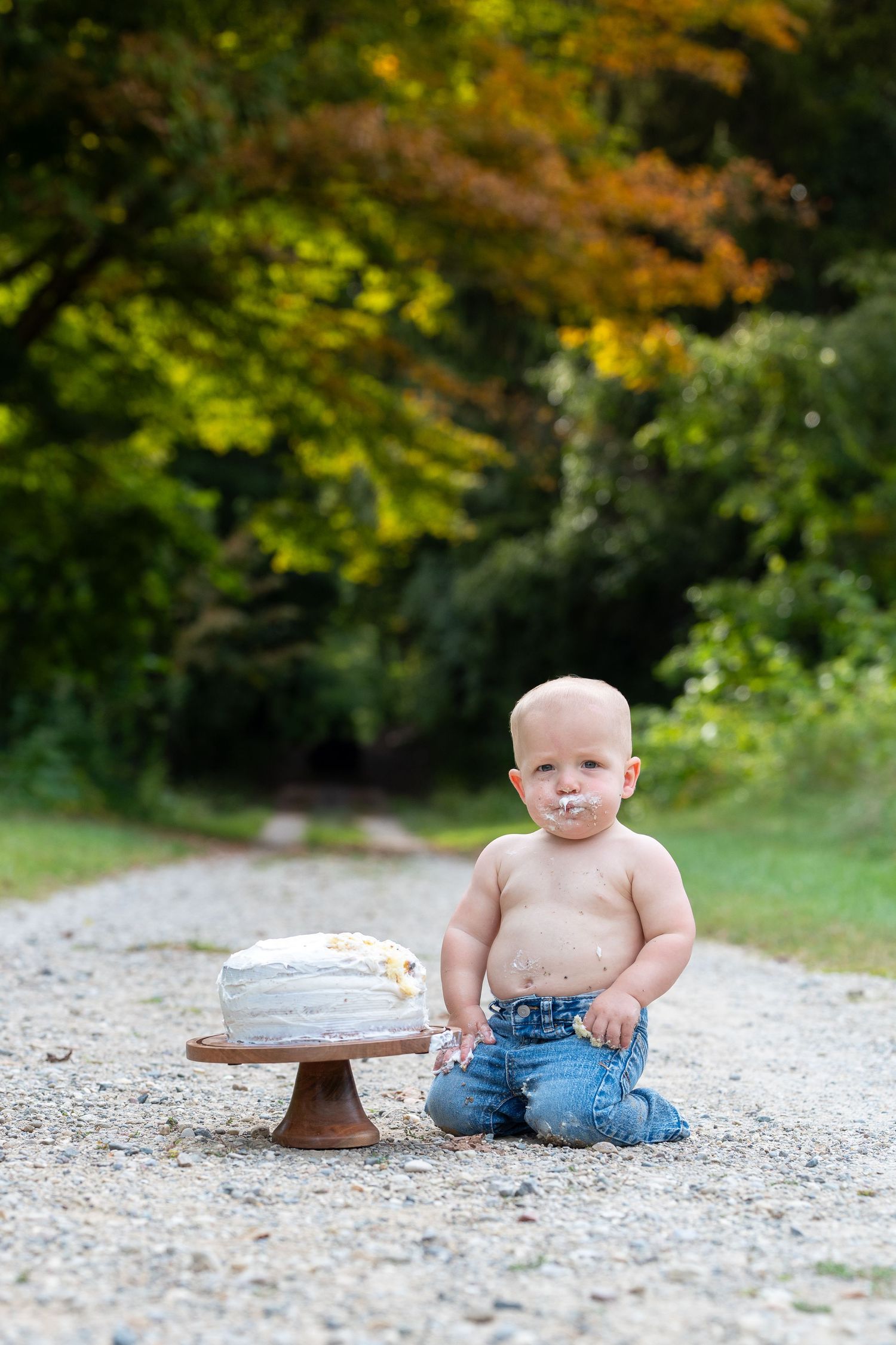Baby wearing jeans sits on gravel path next to small wooden toy in autumn woodland setting.