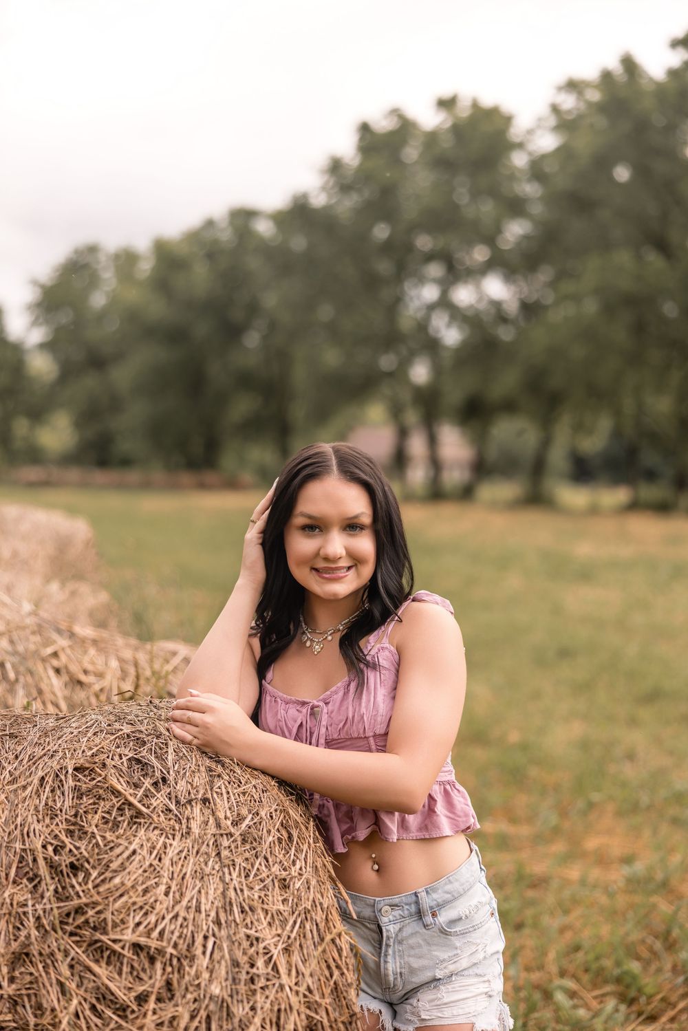 Person in pink tank top and denim shorts poses casually next to a hay bale in a rural field.