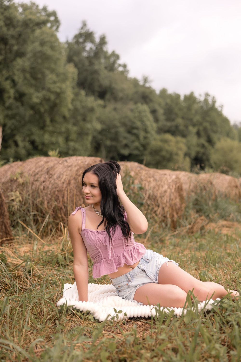 Rural portrait session with model in pink top and denim shorts reclining on white blanket near hay bale.