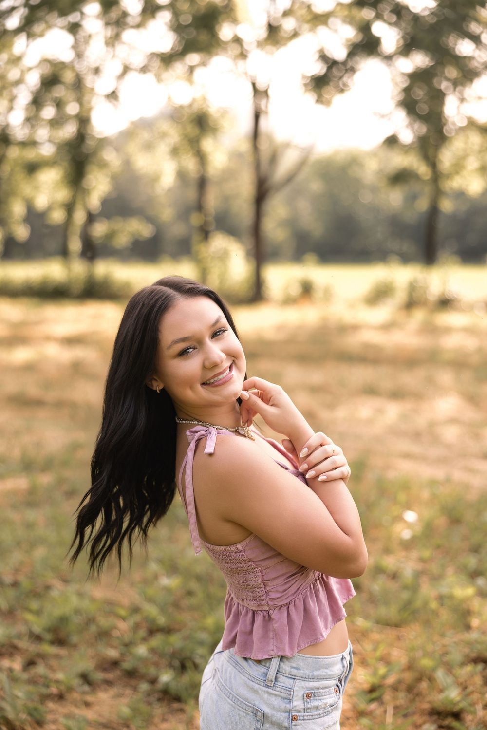 A person wearing a pink top and denim shorts twirls playfully in a sunlit outdoor setting.