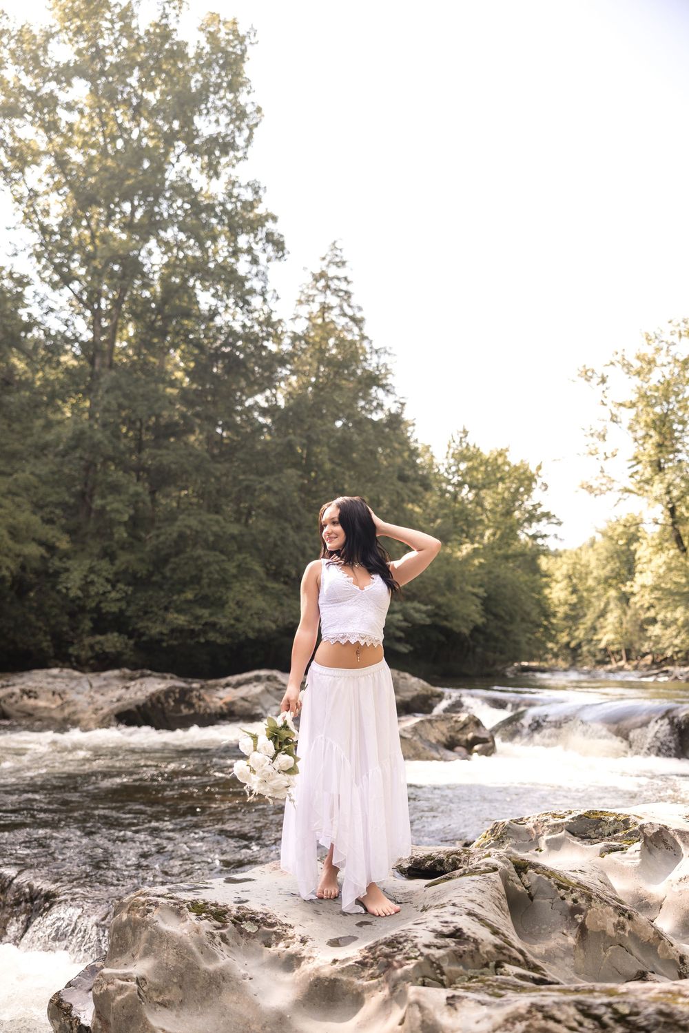 A person in a flowing white dress stands gracefully on rocky terrain near a rushing waterfall.