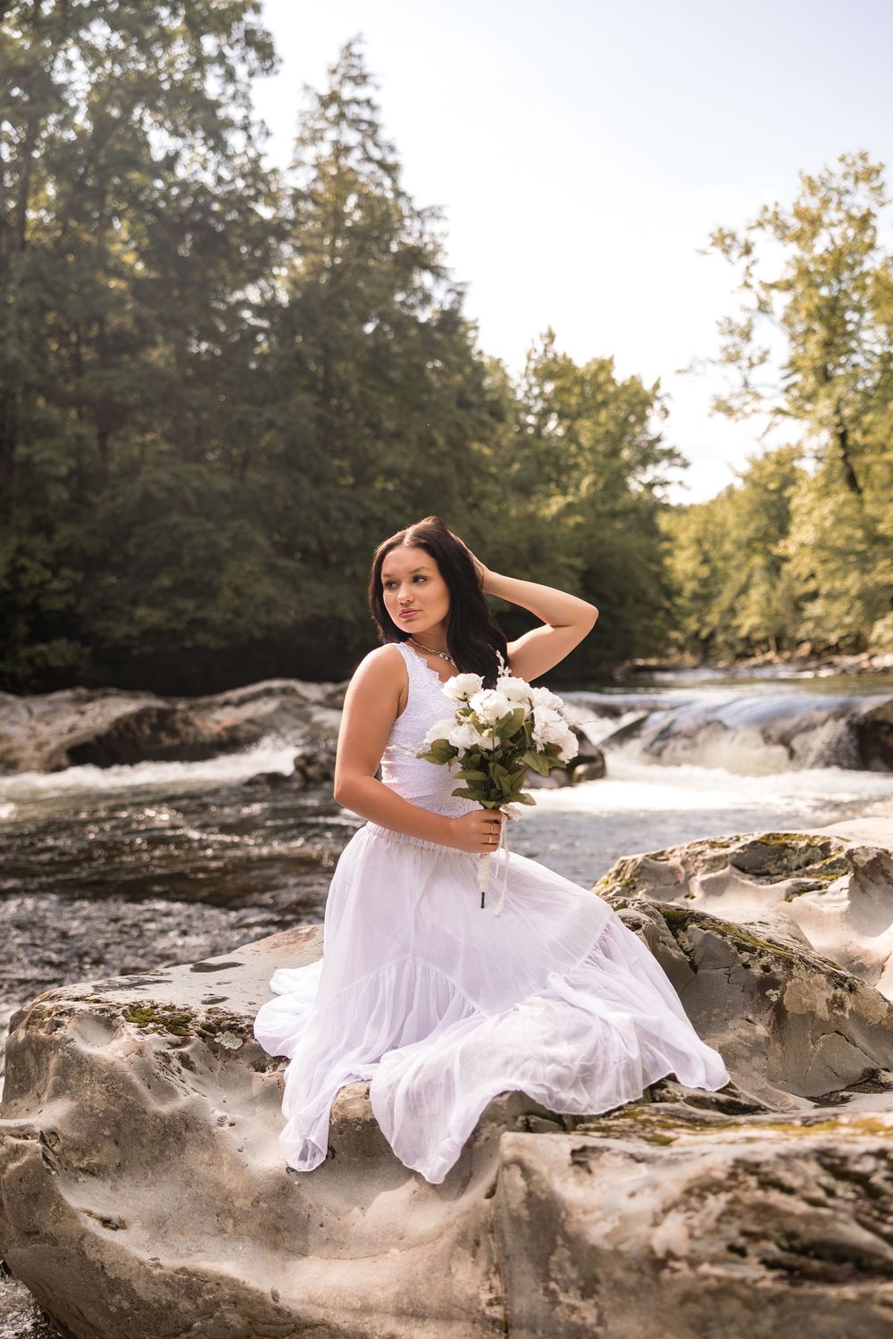 A person in white dress poses elegantly on rock formations by a flowing stream at sunset.