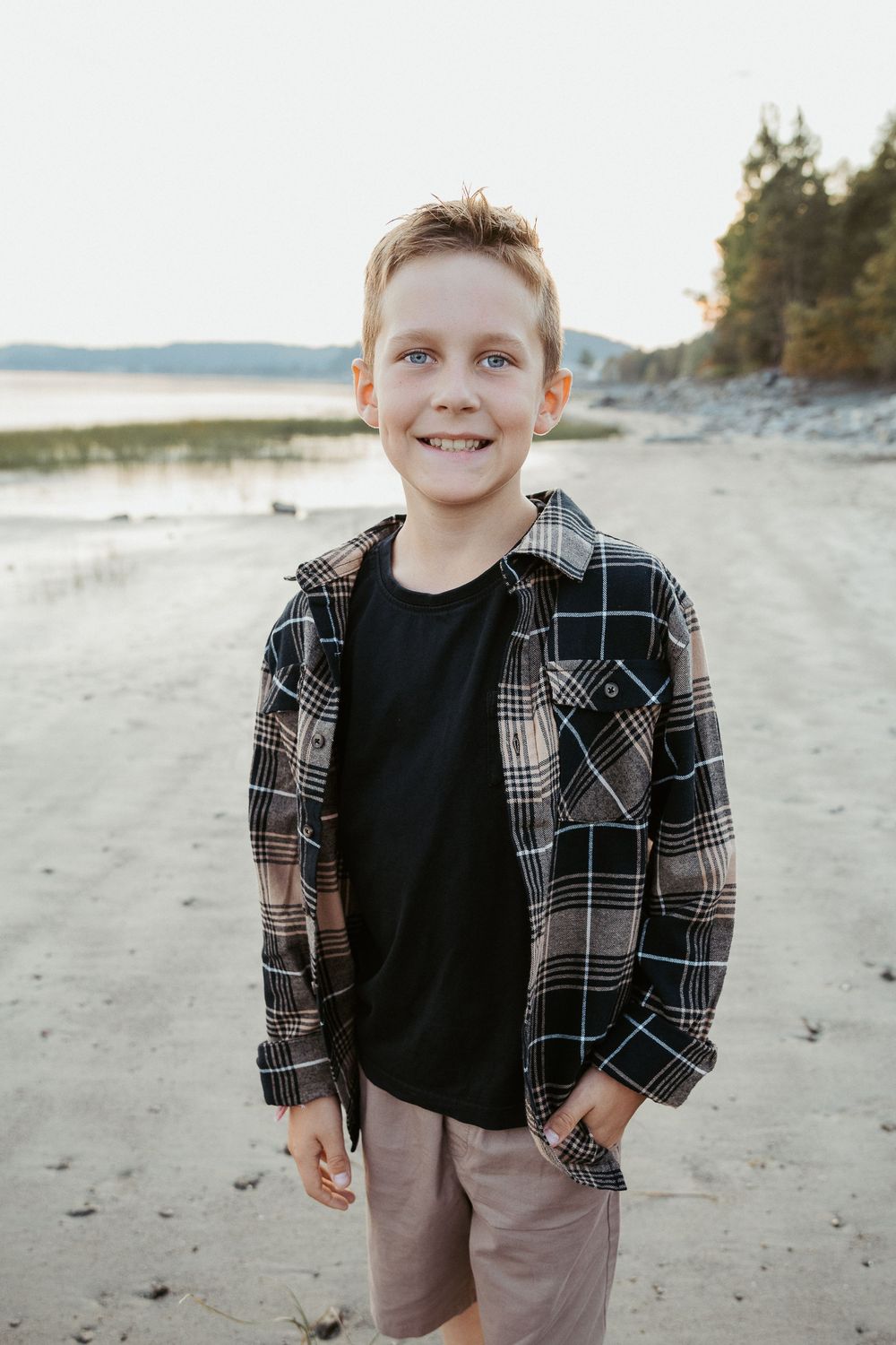 Young person wearing plaid flannel and t-shirt poses on beach with water in background.