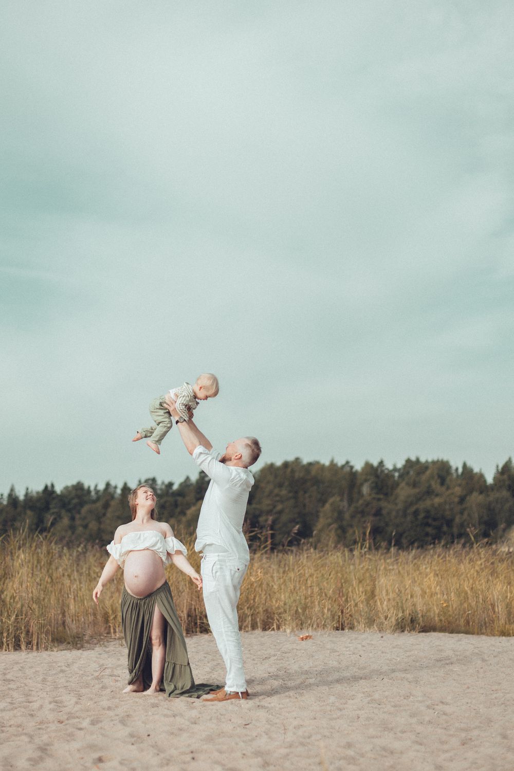 Father lifting baby high in air next to pregnant woman on sandy beach with forest background. Odotuskuvaus koko perheen kera miljöössä.