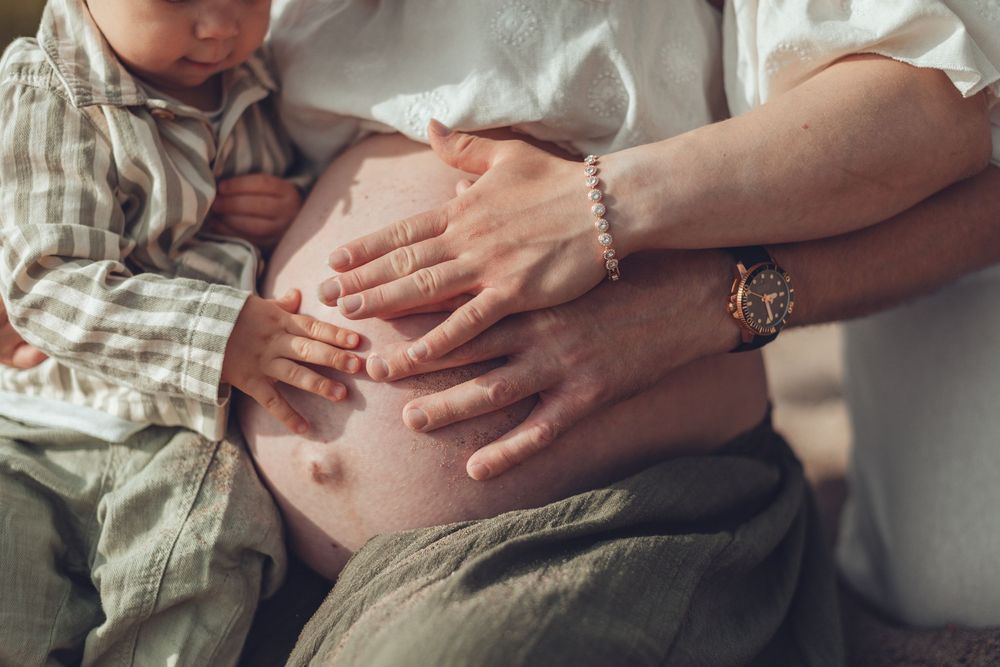 Close-up of pregnant belly with toddler and adult hands gently resting on it outdoors. Odotuskuvaus miljöössä.