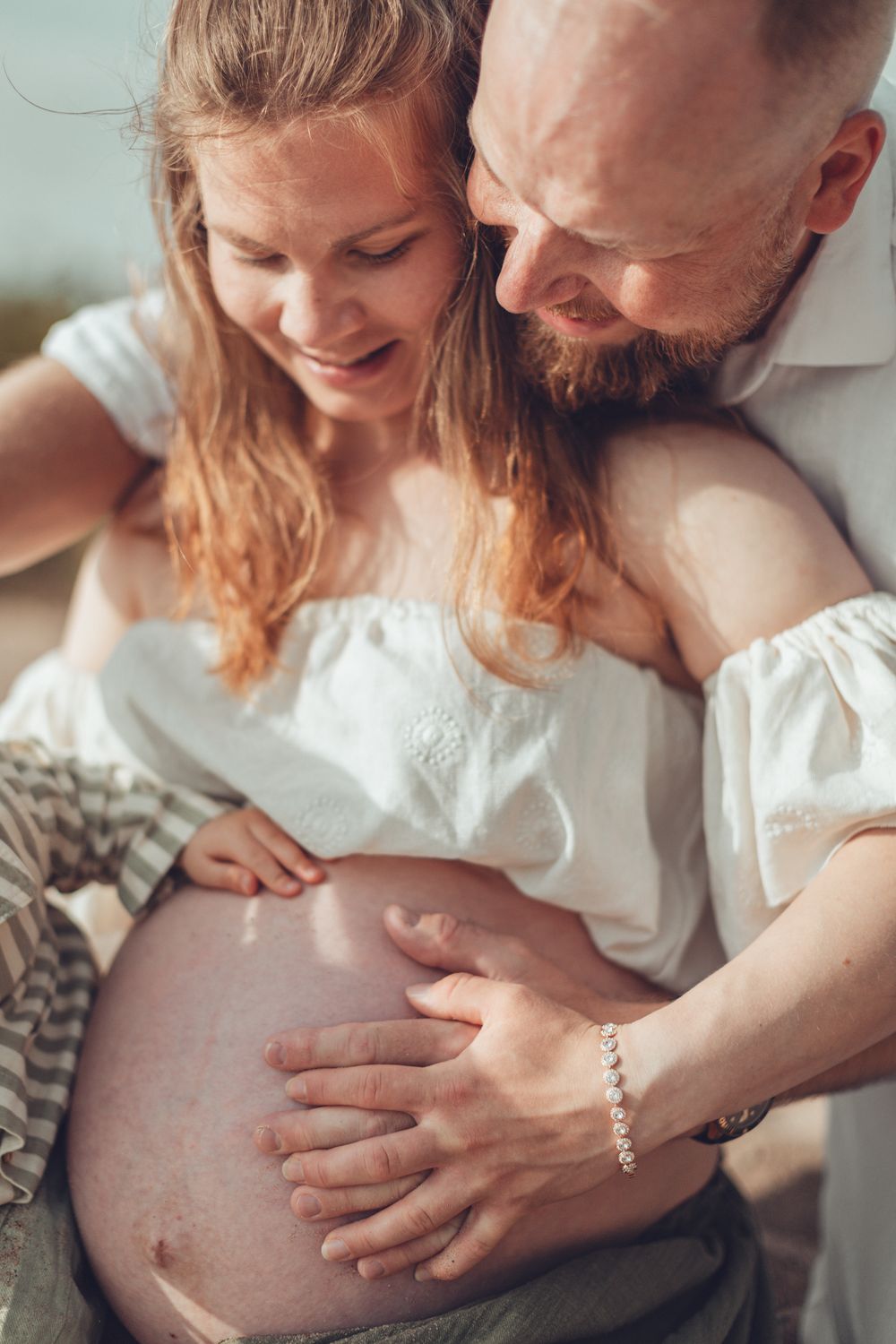 Bearded man embracing smiling pregnant woman from behind, both touching her bare belly outdoors. Odotuskuvaus miljöössä.