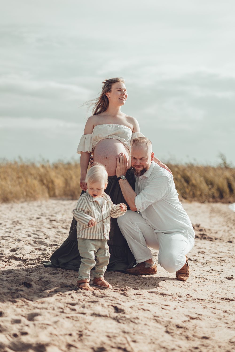 Pregnant mother and bearded father playing with their toddler son on a sandy beach outdoors.