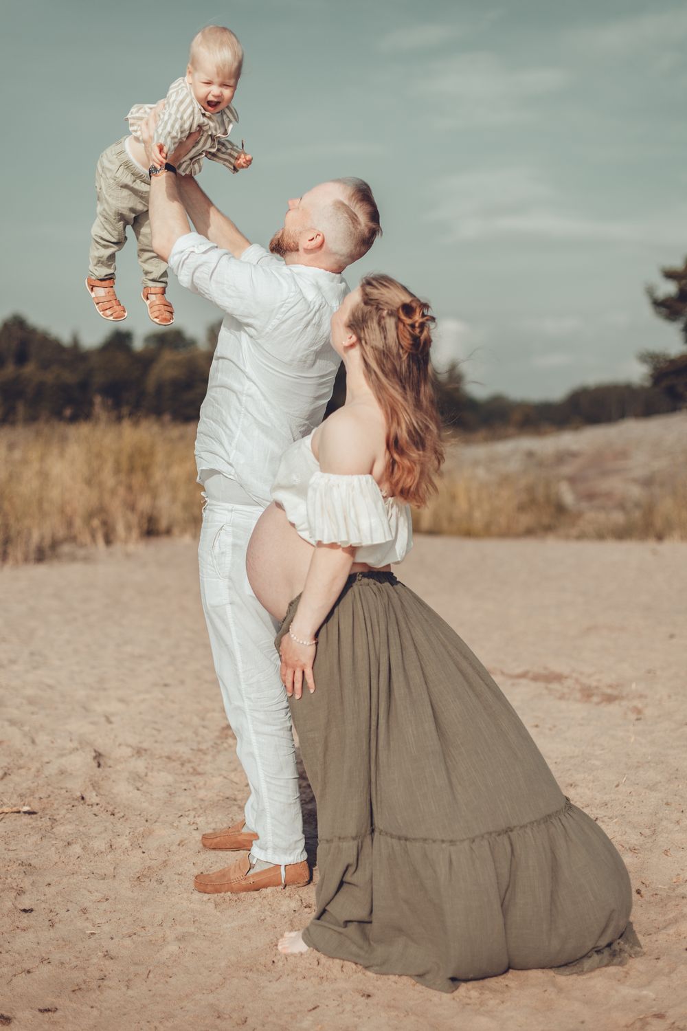Joyful father lifting laughing toddler son while pregnant mother stands beside dad on sandy beach smiling towards toddler.