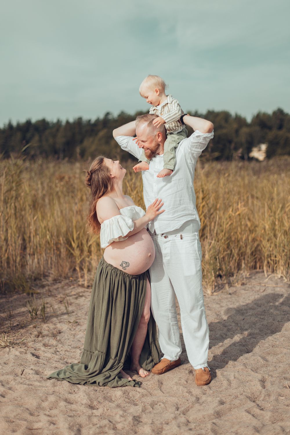 Pregnant woman in white top and green skirt with blond haire husband holding toddler on shoulders on sandy beach. Perhekuvaus, odotuskuvaus.