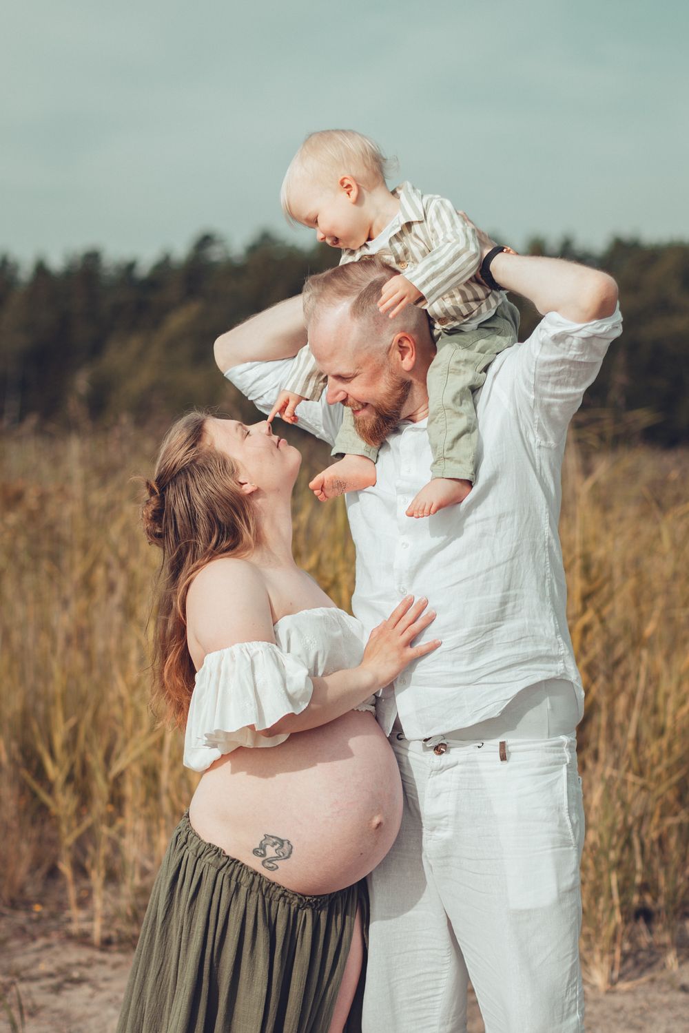 Pregnant woman and husband smiling as their toddler son sits on dads shoulders at the beach. Perhekuvaus, odotuskuvaus.